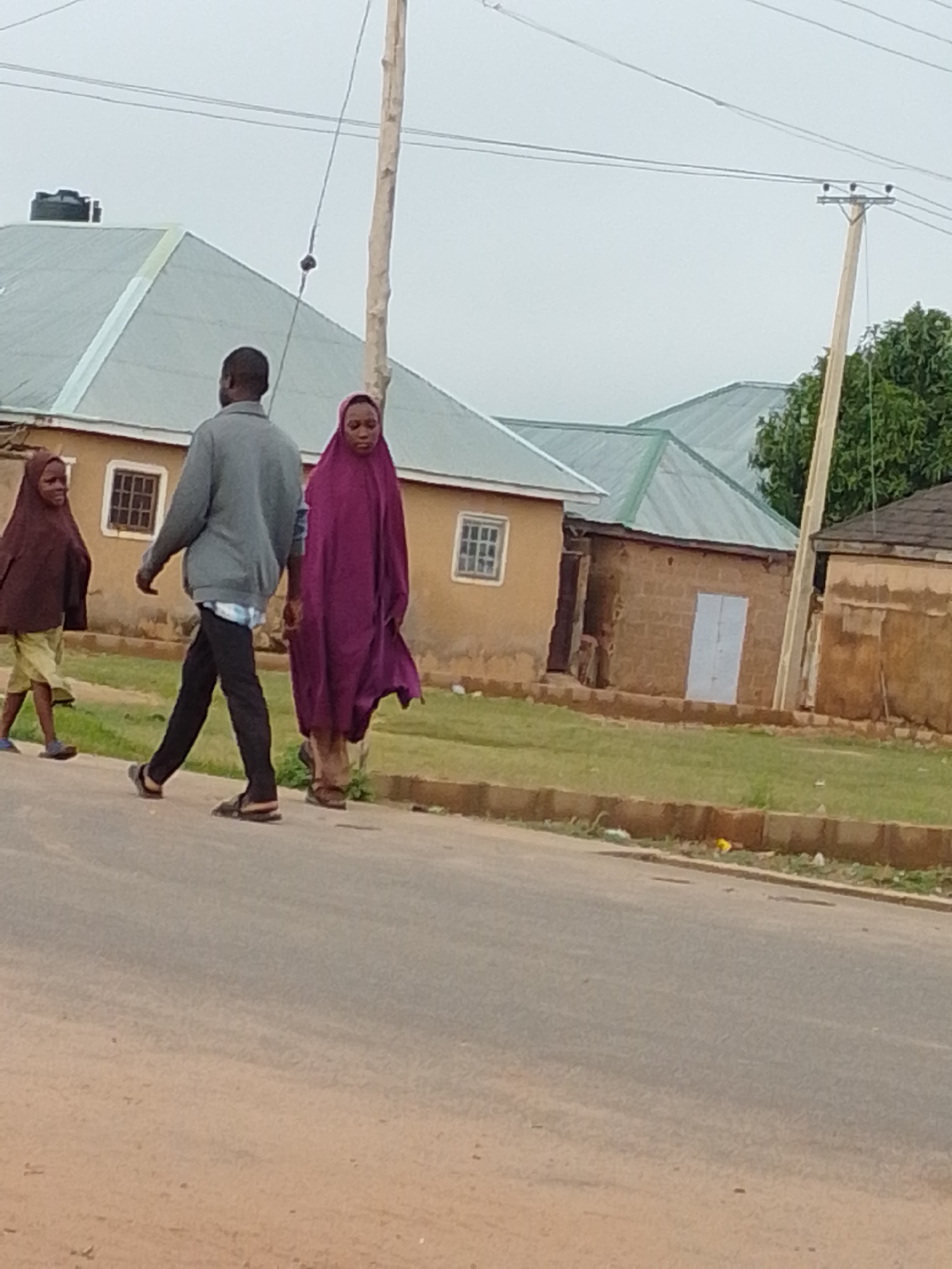 In the city of Unknown, Unknown, three individuals are walking on a paved road, with buildings and a utility pole visible in the background. A person in a gray long-sleeved shirt and black pants, wearing sandals, walks in the center. To their left, a child wearing a brown hijab and light-colored shorts, also in sandals, walks ahead. To the right of the person in gray, a woman in a purple hijab and long dress walks behind. The buildings have light-colored walls and green roofs, with some showing brickwork. A wooden utility pole with wires stands to the right of the scene. The sky is overcast.