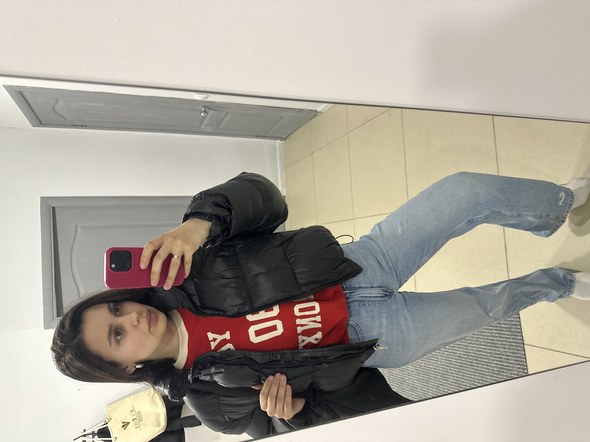 A young woman is captured in a mirror selfie, standing casually indoors. She wears a black puffer jacket over a red long-sleeved shirt, light blue jeans, and white socks. She holds a pink smartphone in her right hand, positioned to take the photo, and a silver ring is visible on her ring finger. Her dark hair is parted to the side, and she looks towards the camera with a neutral expression.
The setting is an interior space, likely a hallway or entrance area in Chaikovsky, Russia. The floor is covered in light-colored tiles, and a gray and white striped doormat is visible beneath her feet. The walls are white, with gray door frames and doors reflected in the background. Above, a white ceiling features a rectangular gray access panel.
Notable details include a beige tote bag with a partially visible, reversed logo and text that appears to read "VXIR YM" hanging to her left. The reversed white text on her red shirt can be interpreted as "MOXY" and "00". The overall impression is a casual outfit documented in a well-lit, everyday indoor environment.