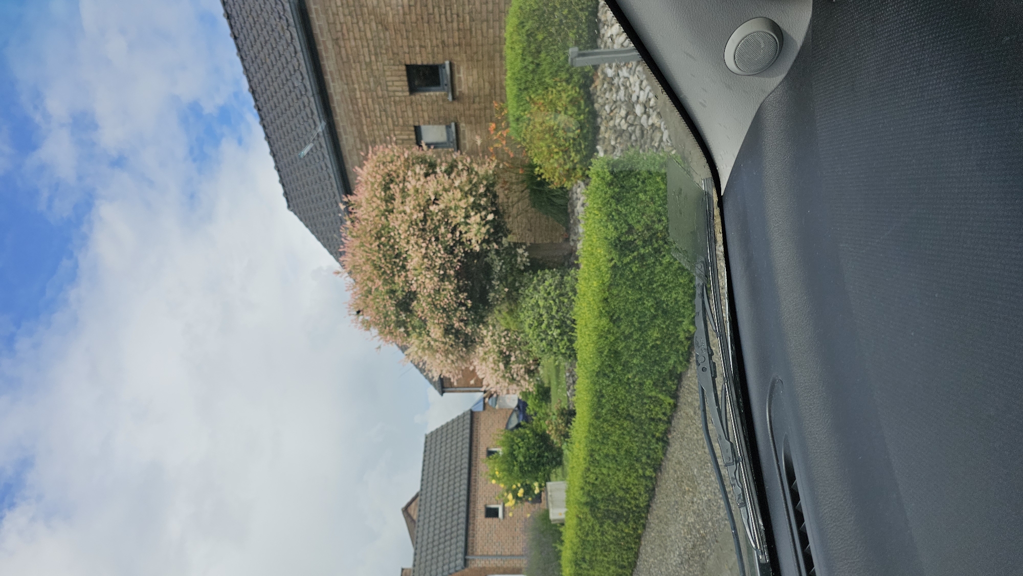The image presents a residential street scene, captured from the interior of a car, looking out through the windshield. The perspective suggests the vehicle is parked or driving slowly along a paved surface, part of which is visible at the bottom of the frame next to a manicured green hedge.
In the background, under a bright blue sky dotted with scattered white clouds, are several brick houses with dark gray tiled roofs. The closest house features dark, rectangular windows and is partially obscured by lush vegetation. A prominent tree with distinctive light pink and green variegated foliage stands in front of this house, adding a vibrant touch of color to the scene. Various other green hedges and shrubs line the properties, indicating well-maintained gardens.
The right side and bottom portion of the image reveal parts of the car's interior: a dark gray textured dashboard, a round speaker unit positioned near the windshield's edge on what appears to be the passenger side A-pillar, and the black wiper blades resting on the windshield.
The time of day is clearly daytime, with ample sunlight illuminating the scene. The weather appears pleasant and clear. There is no visible text in the image. The architectural style of the houses and the general landscape are consistent with a suburban or rural residential area, such as in Thuin, Belgium.