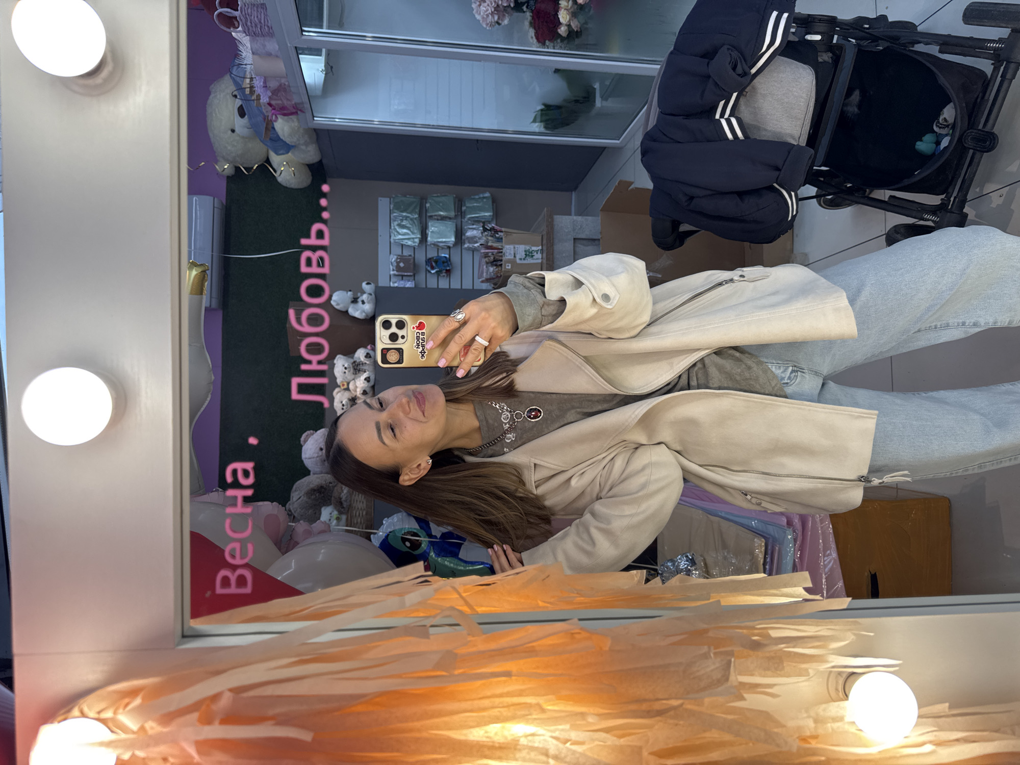 A woman takes a selfie in front of a mirror adorned with lights and streamers. Reflected in the mirror are flower arrangements, stuffed animals, balloons, and a sign in Russian that says "Spring, Love". A baby stroller is visible in the background. The image was taken in Sochi, Russia.