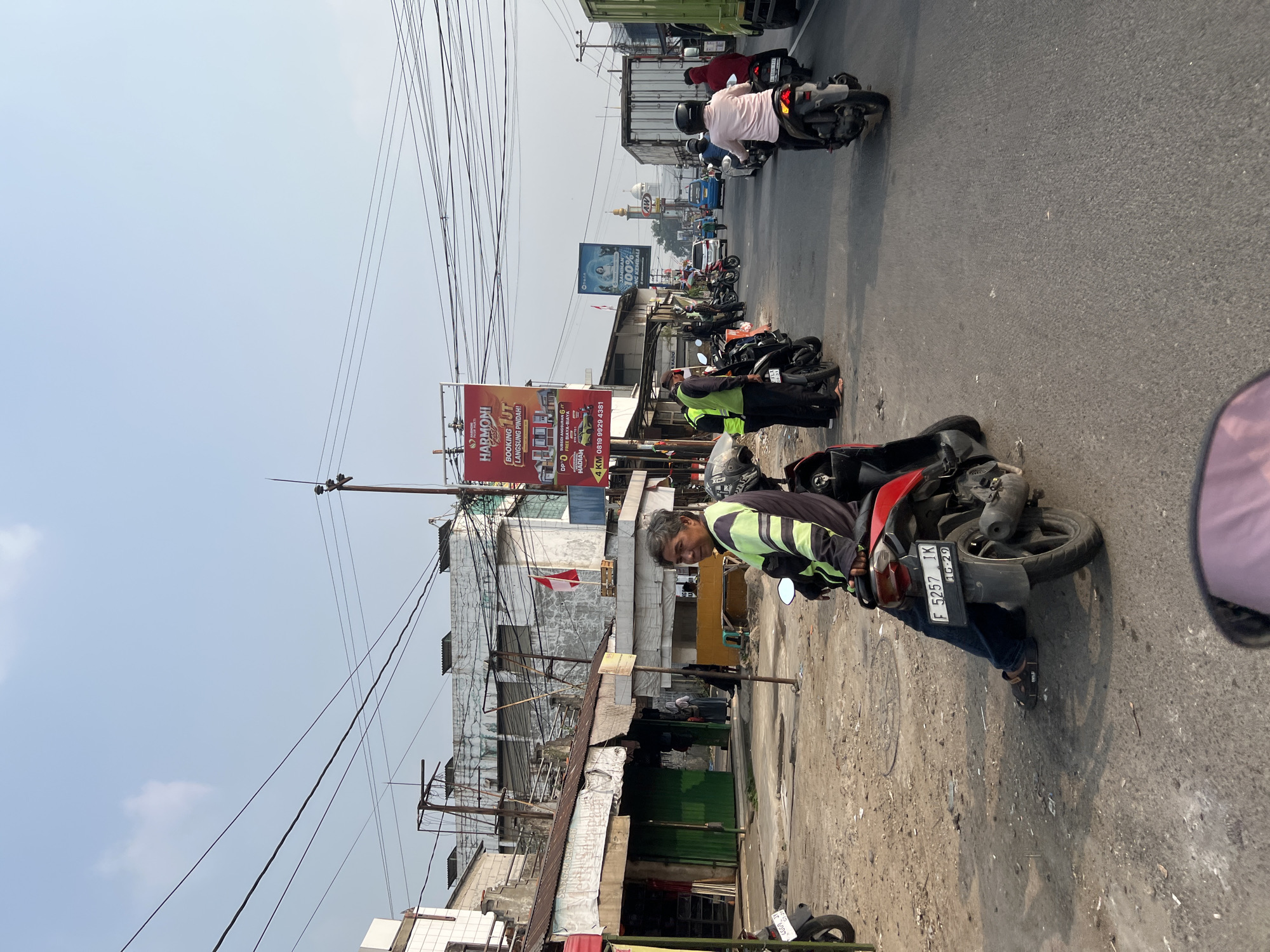The image captures a street scene in Citeureup, Indonesia, characterized by traffic congestion and local commerce. In the foreground, a man sits on a red motorcycle, wearing a reflective green and black jacket and dark blue jeans. The motorcycle's license plate reads "F 5257 IK". Beside him, another motorcycle with license plate "4409 JJ" is parked. The road is paved with asphalt, and the surrounding area is a mix of dirt and gravel.
To the left, a row of small storefronts with corrugated metal roofs lines the street. Some of these shops have green metal shutters, and a red sign is visible on the left edge of the frame. A few people are standing or walking near these shops. In the background, buildings with similar corrugated roofs are visible, along with utility poles and a network of overhead wires.
Further down the street, traffic is moderate, with several other motorcycles and a few cars. A white truck is prominent on the right side of the road. Two large billboards are displayed prominently. One, a red billboard, advertises "HARMONI" and provides a phone number. Another blue billboard promotes a service with the text "JAMINAN 100%". A mosque with a dome and minarets is visible in the distance, suggesting the cultural context of the location. The sky is clear and blue, indicating a sunny day. The overall impression is a bustling, everyday scene in a semi-urban Indonesian environment.