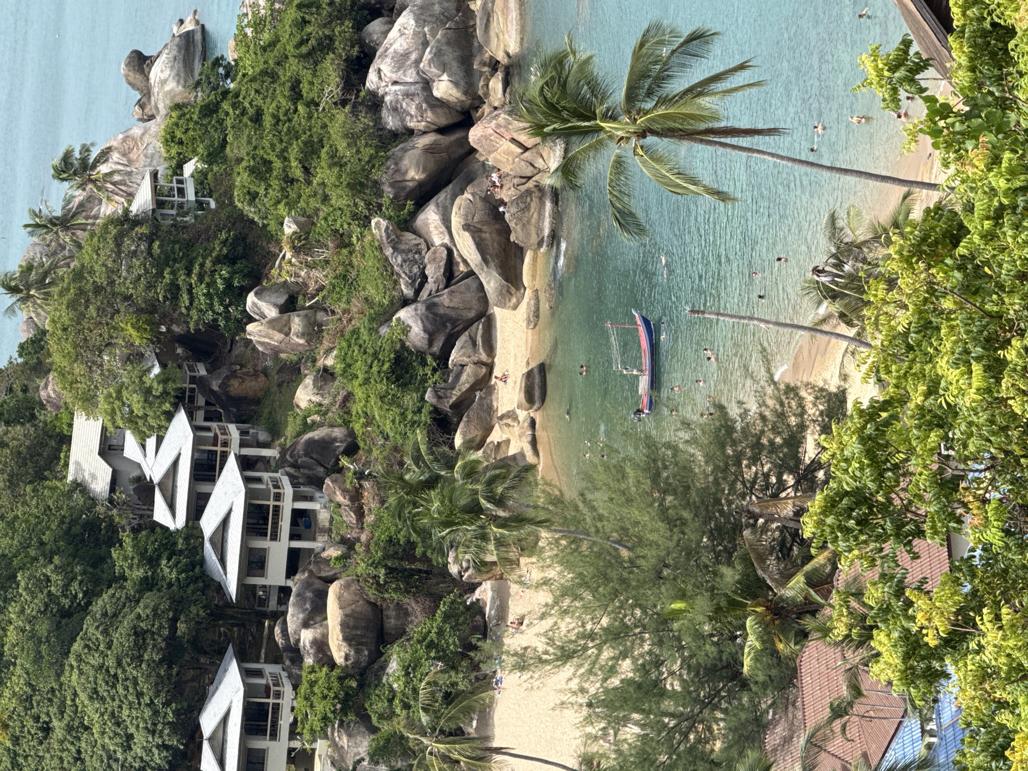 An aerial view of a tropical cove in Koh Samui District, Thailand, reveals a secluded beach dotted with sunbathers and swimmers. Nestled on the lush, rocky hillside overlooking the cove are several modern villas with white facades and gray roofs, their balconies offering panoramic views of the turquoise waters. The scene is further enhanced by the presence of a small fishing boat anchored in the bay and lush green vegetation, including palm trees, that cascades down the slopes towards the sandy shore. The water is clear and shallow, inviting people to swim and cool off under the tropical sun.