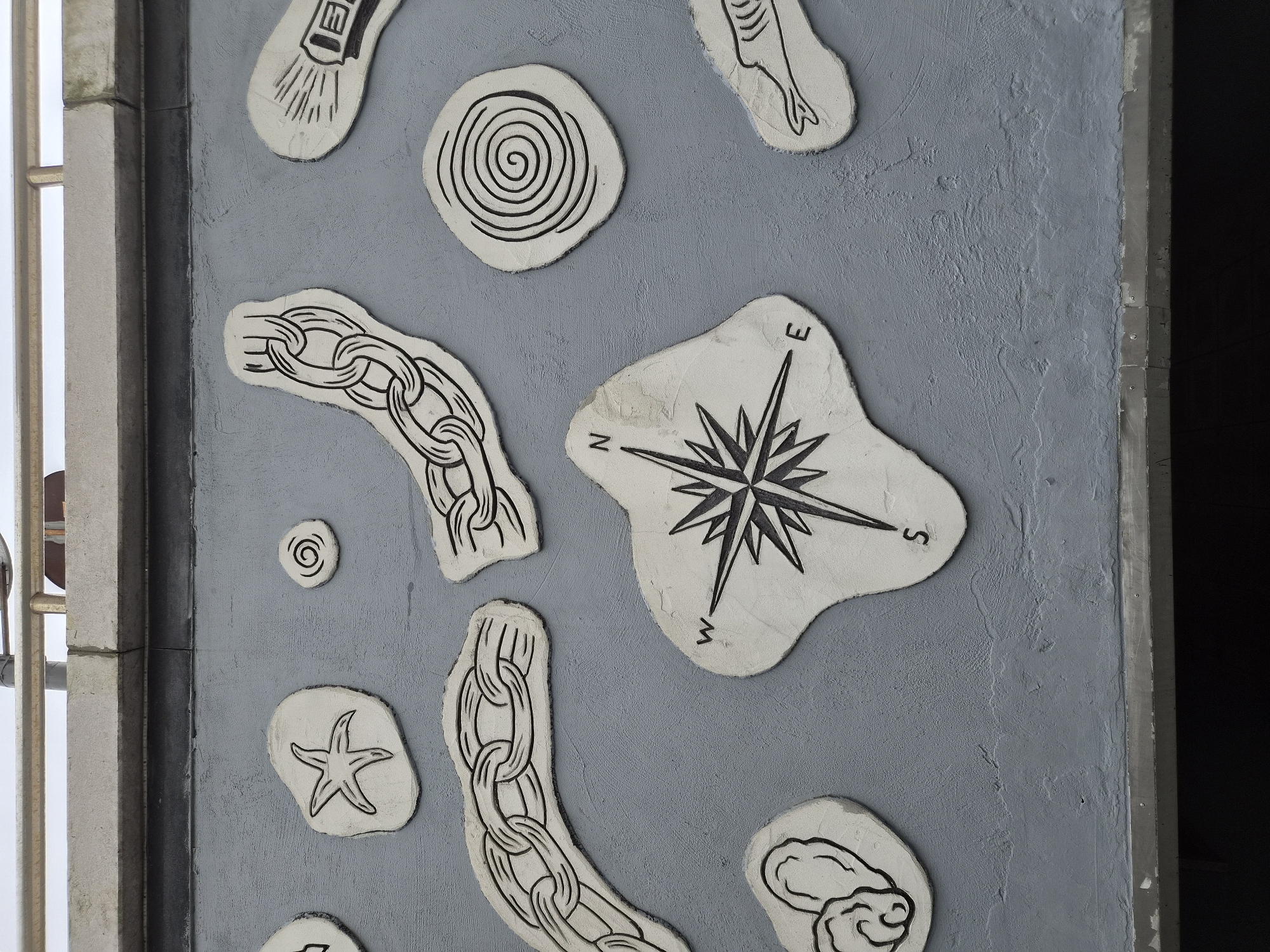 The image shows a textured grey wall decorated with white relief sculptures outlined in black. The sculptures depict various nautical and seaside themes. Prominently featured is a compass rose in the center, with the letters "N," "E," and "S" visible. To the left of the compass, a starfish and a spiral shell are depicted. Above the compass, two segments of a chain are shown, one larger and more detailed than the other. To the right of the compass, a spiral shape and what appears to be a drawing of a fish are present. The wall itself has a rough, concrete-like texture and is located outdoors, with a metallic railing and a grey ledge visible at the top of the frame. The bottom of the image shows a dark, out-of-focus area, likely a walkway or structure below. This art installation is located on a wall in Vladivostok, Russia.