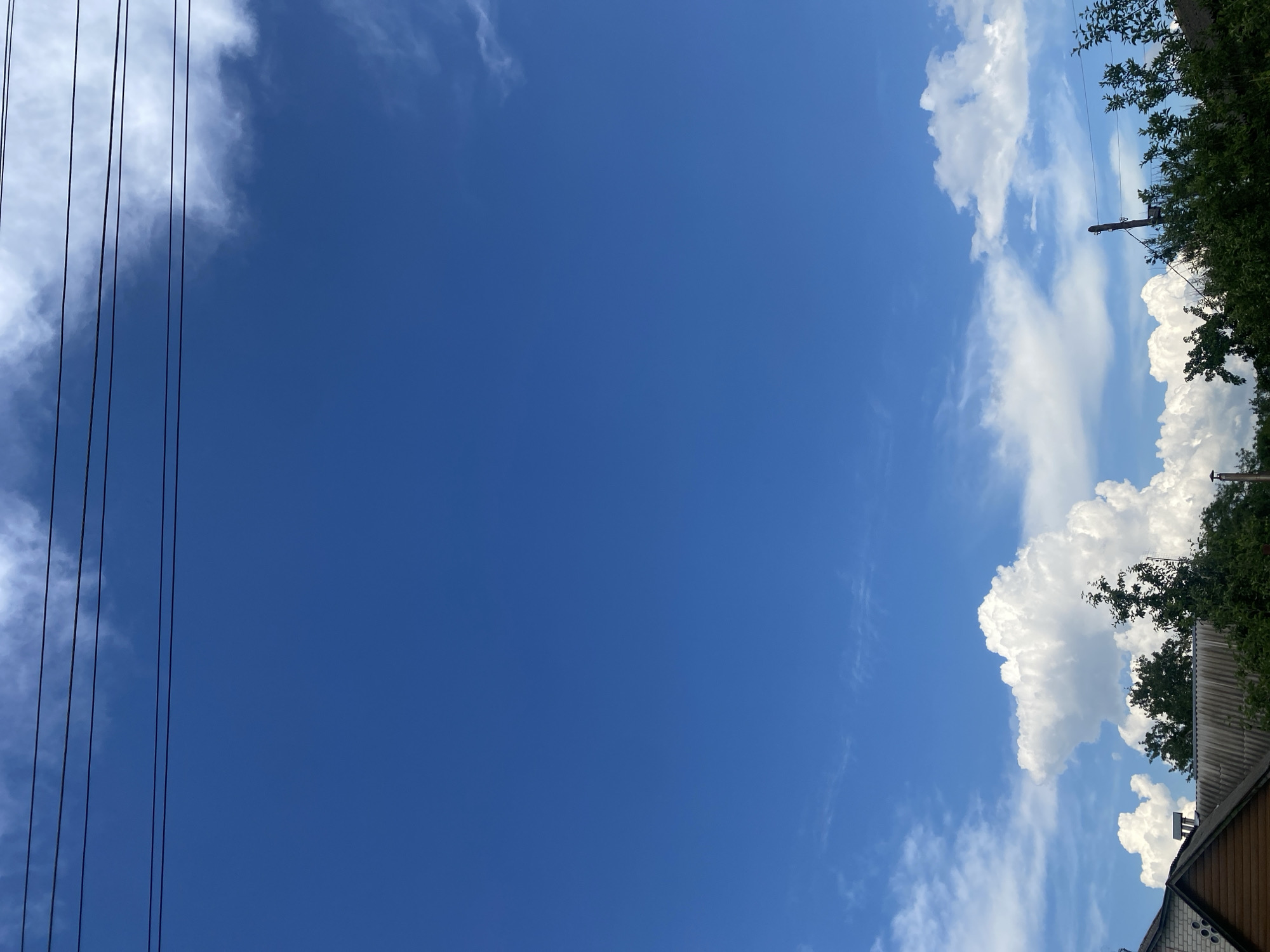 The image depicts a clear blue sky with scattered white clouds above a residential area. Power lines are visible in the upper portion of the frame. In the foreground, the rooftops of houses and dense green trees can be seen. The architecture suggests a rural or suburban setting. The scene is peaceful and shows a typical day in Chernihiv, Ukraine. There are no people or vehicles visible in the image. The lighting indicates daytime.