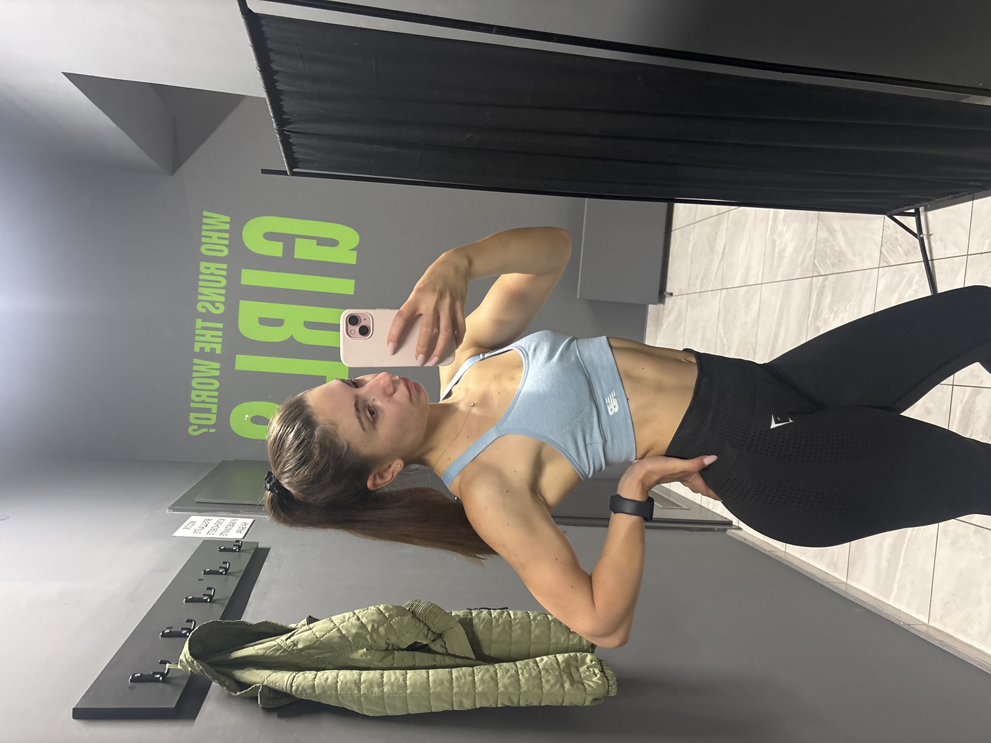A young woman with brown hair in a ponytail, wearing a light blue sports bra and black leggings, stands in a locker room, taking a mirror selfie with her phone. The locker room has grey walls and a tiled floor. A green quilted jacket hangs on a wall-mounted rack to the left of the woman. In the background, neon green text on the wall reads "WHO RUNS THE WORLD? GIRLS". A black changing room curtain is visible to the right. The location is the city of Varna, Bulgaria.
