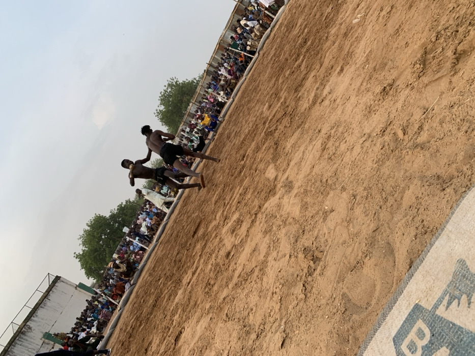 The image shows a wrestling match taking place on a sandy field in Sokoto, Nigeria, located at latitude 13.049 and longitude 5.238. Two men, shirtless, are engaged in a wrestling match. They are in the middle of the field, surrounded by a crowd of spectators. The spectators are seated on the perimeter of the field, with a white building in the background. The men are both wearing shorts and appear to be of African descent. One man is wearing a black pair of shorts while the other is wearing a pair of patterned shorts. The image is taken from a low angle, looking up at the wrestlers. The sand is brown and dry.