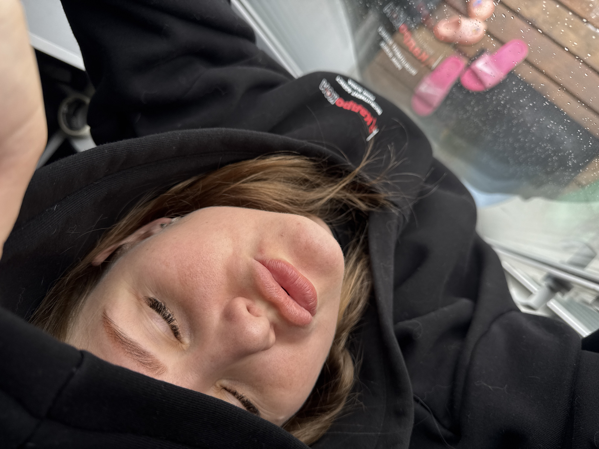 A close-up, slightly angled shot shows a person wearing a black hooded sweatshirt. The person's eyes are mostly closed and the face is positioned towards the camera. The background includes a blurred window with raindrops, revealing a glimpse of pink sandals and a wooden surface, possibly a deck or patio. Part of the hood obscures the face. A logo is visible on the black sweatshirt, but the details are unclear. The environment appears to be indoors, looking out through a window. The location in the city of Gornyi (Dvurechenskoe S/P), Russia, cannot be determined from the visual information alone.