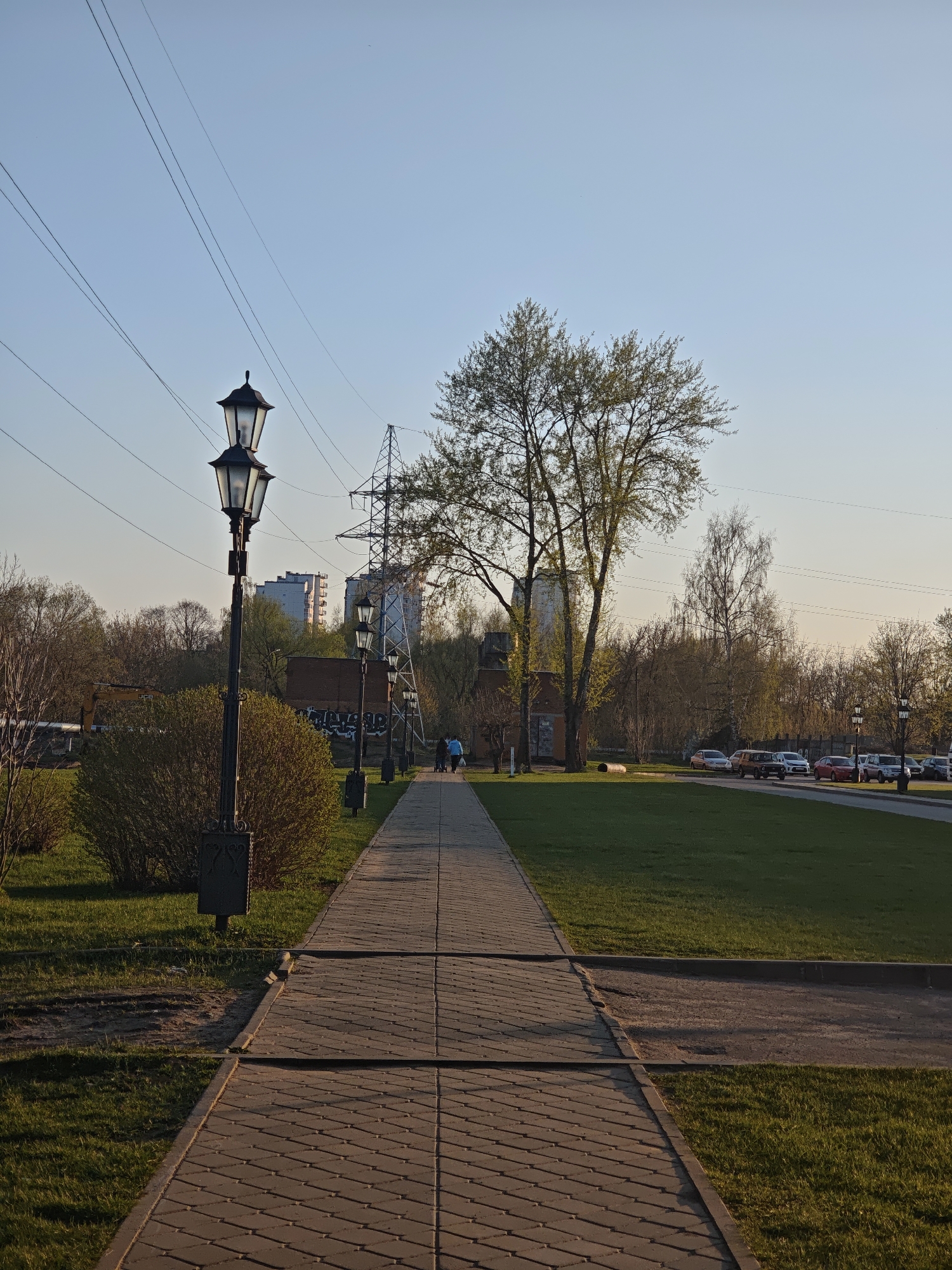 The image captures a pathway in a park, presumably in Noginsk, Russia, as suggested by the lighting and architectural style. The path, paved with interlocking blocks, leads straight into the distance, framed by manicured grass lawns on either side. Along the left side of the path, there are several ornate street lamps with multiple lanterns. Bushes and trees line the left side, with buildings visible further back. An electrical transmission tower stands in the middle ground, suggesting proximity to power lines that run across the top of the frame. In the distance, several parked cars are visible on the right side. The sky is a clear, pale blue, indicating either early morning or late afternoon. Two individuals, possibly a parent with a child in a stroller, are faintly visible walking down the path.