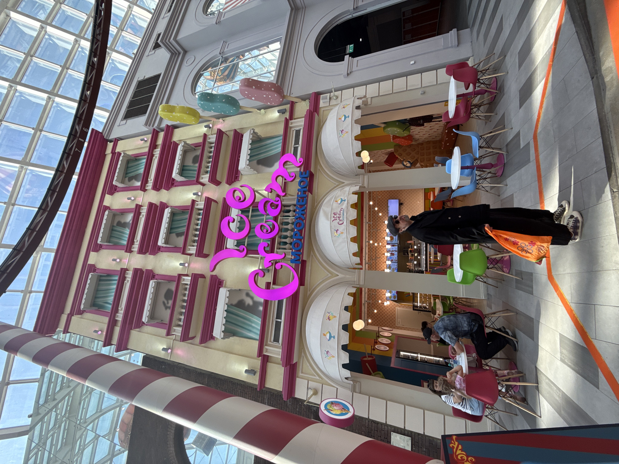 The image depicts the interior of a brightly colored, stylized building, identified by the Russian word "Mopoжeнoe" (Ice Cream) in large, magenta letters. The building facade is multi-storied with pink and white trim and pastel-colored lollipop decorations. Below the facade are awnings with an ice cream cone design, beneath which patrons are seated at tables.
A man dressed in black stands in the aisle. Another man and two children are seated at a table, interacting.
A large candy cane-striped pole ascends into the ceiling, which features a grid-like glass structure. The location is in Moscow, Russia, as indicated by the Cyrillic text on the building.