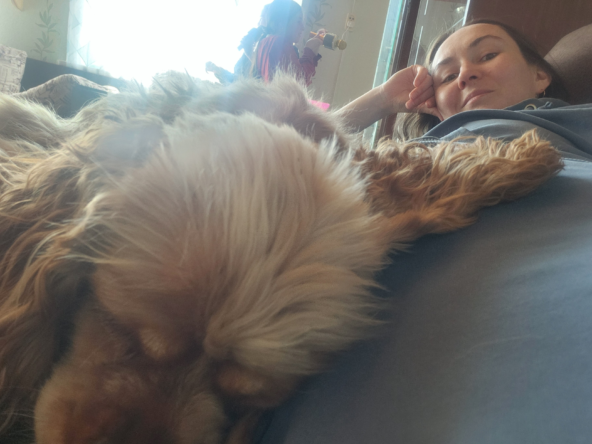 A woman with shoulder-length brown hair lies on her back, resting her head on her hand, with a brown and white dog lying on her chest. The background is blurred, but shows a window, curtains, and a person holding a microphone. The scene appears to be indoors. The location is Архангельское, Russia.