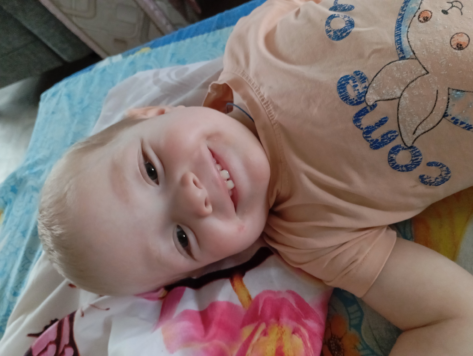 A young child lies on a patterned bed covering in Yurgamysh, Russia. The child, with fair skin and short blonde hair, is smiling and facing the camera. The child is wearing a peach-colored t-shirt with a blue printed design, featuring the word "COME" and a unicorn. The bed covering has a floral pattern in pink and yellow. The background includes a section of a blue and white patterned blanket, and a hint of a dark-colored piece of furniture.