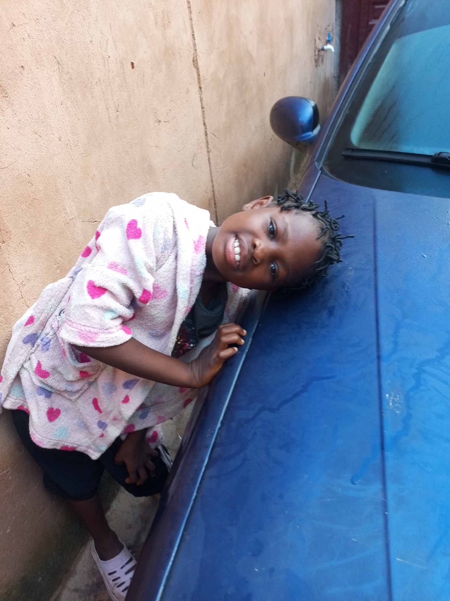 A young Black girl with braided hair, wearing a pink and white robe with heart patterns, lies on the hood of a blue car. She is leaning against a textured, tan wall and smiles at the camera. Her feet are clad in white sandals. The scene appears to be outdoors, possibly in an alleyway or residential area within Tempure, Nigeria, given the visible wall, car, and outdoor setting.