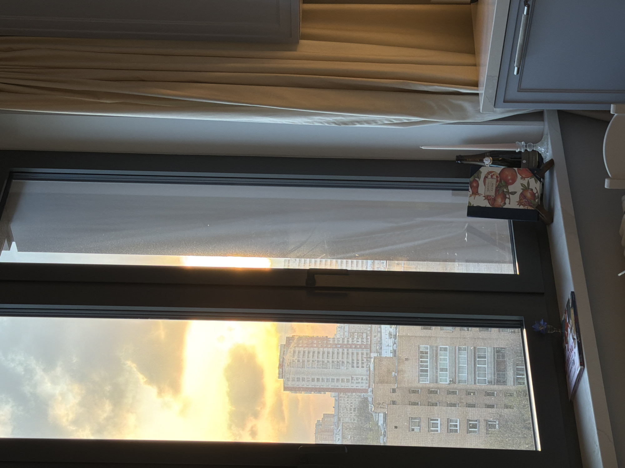 The image presents an interior view looking out through a large window onto an urban landscape, likely at either sunrise or sunset due to the prominent golden light. Inside, light beige curtains are drawn back at the top of the window, above a white roller blind that partially covers the upper window pane, through which a bright light source is visible.
On the right side of the window sill, several objects are arranged: a tall, dark bottle, a clear glass candlestick holding a light pink taper candle, and a decorative book or box adorned with an illustration of pomegranates. A portion of a grey kitchen cabinet with a silver handle is visible above these items, and in the lower right corner, a small vase containing a single blue flower is tucked against the window frame.
The exterior view reveals a cityscape dominated by numerous multi-story buildings, their facades reflecting the warm, golden light that floods the sky. Large clouds, illuminated by the low sun, create a dramatic and colorful backdrop. The architecture suggests a dense urban setting, consistent with Moscow, Russia, which is provided as the location context. No people or specific activities are visible. The text on the pomegranate-decorated item is not clearly legible.