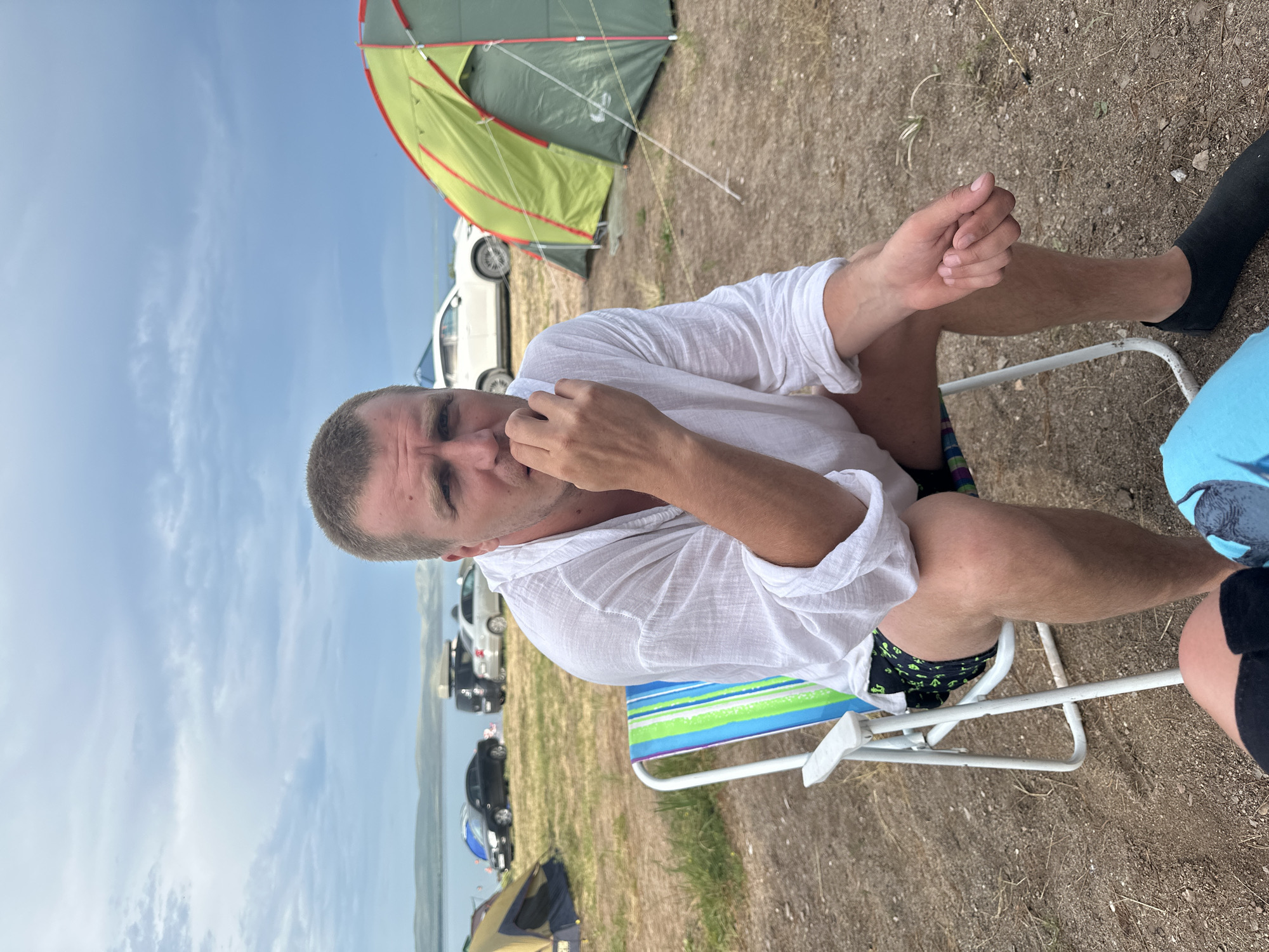 A man is sitting in a striped beach chair outdoors, facing the camera. He is wearing a white, loose-fitting long-sleeved shirt and patterned shorts. His hair is short and brown, and he has a light beard. He is holding his right hand to his mouth, as if eating or scratching. The man has a neutral expression.
Behind him, to his left, a green and yellow tent is partially visible, along with several cars parked in the background. The ground appears to be sandy and dry, with some sparse vegetation. The sky is blue with scattered white clouds, suggesting it is daytime. The overall scene suggests a camping trip or outdoor gathering. The man's attire and the presence of the beach chair and tent indicate a casual, recreational setting, possibly near a body of water given the general appearance of the landscape. The location context provided is Zhemchuzhny, Russia. There is no visible text in the image.