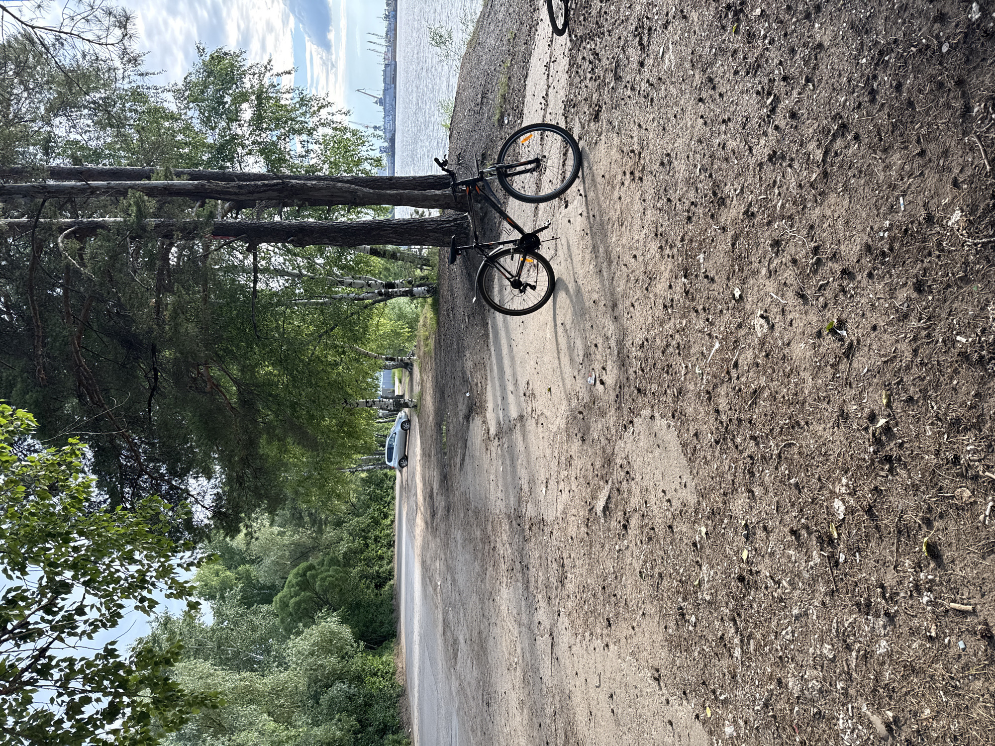 The image depicts a dirt path next to a body of water, likely a river or sea, with industrial cranes visible in the distance. A black bicycle is parked on the path, leaning against its kickstand. The path is lined with trees on the left, creating a sense of a natural, possibly park-like, setting. Further down the path, a white car is visible, and beyond that, more trees and greenery. The sky is partly cloudy, suggesting daytime. The overall mood is tranquil and serene, with the presence of the bicycle and car hinting at human activity in the area. The location appears to be Chaikovsky, Russia, given the context. There is no visible text in the image.