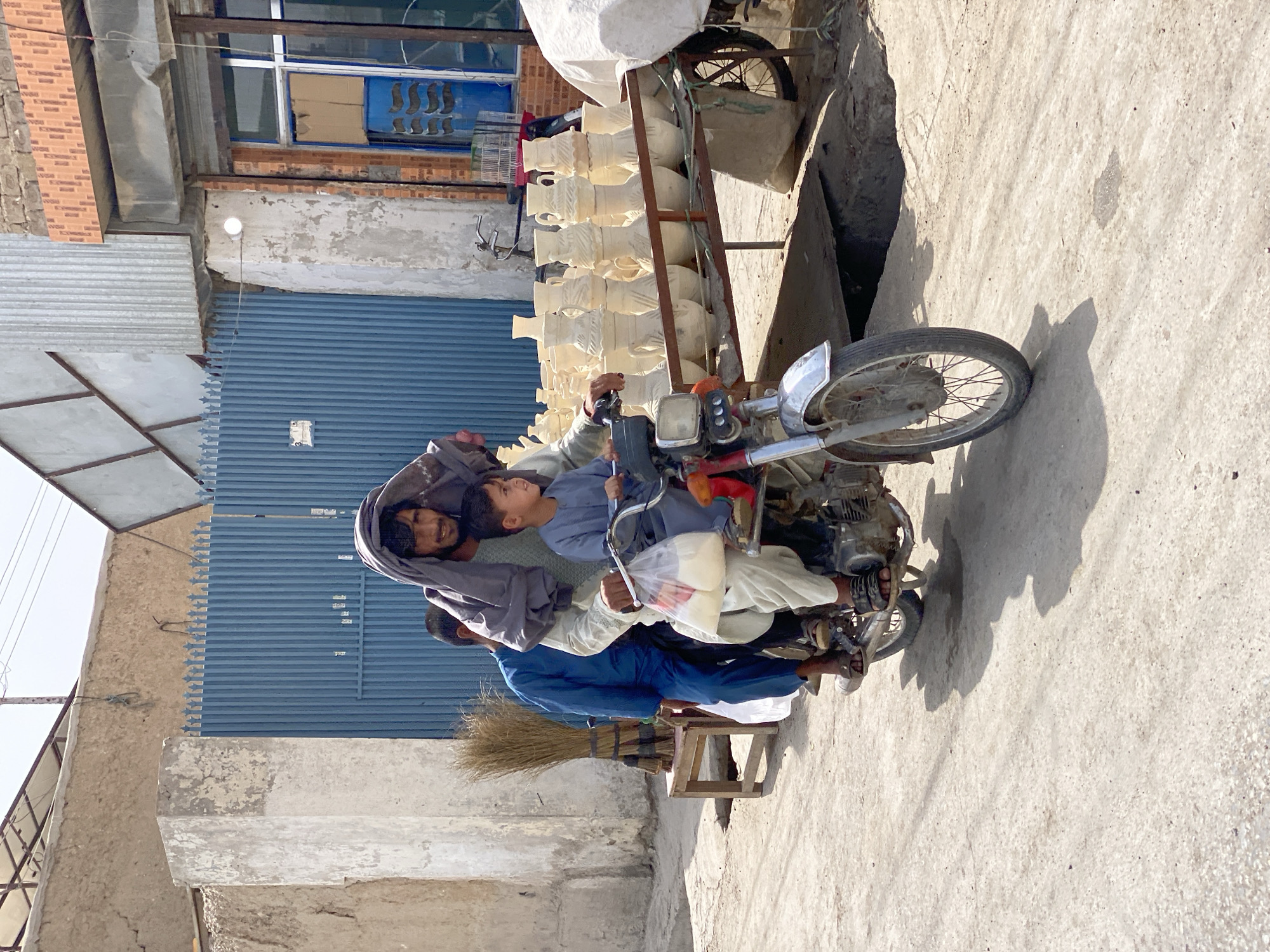 In Kandahar, Afghanistan, a man with a headscarf draped over his head rides a motorcycle, carrying two passengers. He is wearing a light-colored tunic and trousers, and his face is partially obscured by the scarf. The passenger behind him, wearing a blue tunic and trousers, is seated behind the driver. A young boy in a light blue tunic and trousers is seated at the front, holding a plastic bag filled with white items. The motorcycle is old and appears to be well-used, with a prominent exhaust pipe. In the background, a blue metal gate with vertical slats stands behind a rough-textured wall. To the right of the gate, a building with a brick facade and large windows is visible. In front of the building, a display rack is filled with numerous cream-colored ceramic vases of varying sizes and shapes, some stacked precariously. A broom with dried reeds stands propped up on a small wooden stool to the left of the gate. The scene is set on a dusty, paved road bathed in bright sunlight, casting strong shadows. The overall impression is one of everyday life in a bustling, outdoor market or street in Afghanistan.
