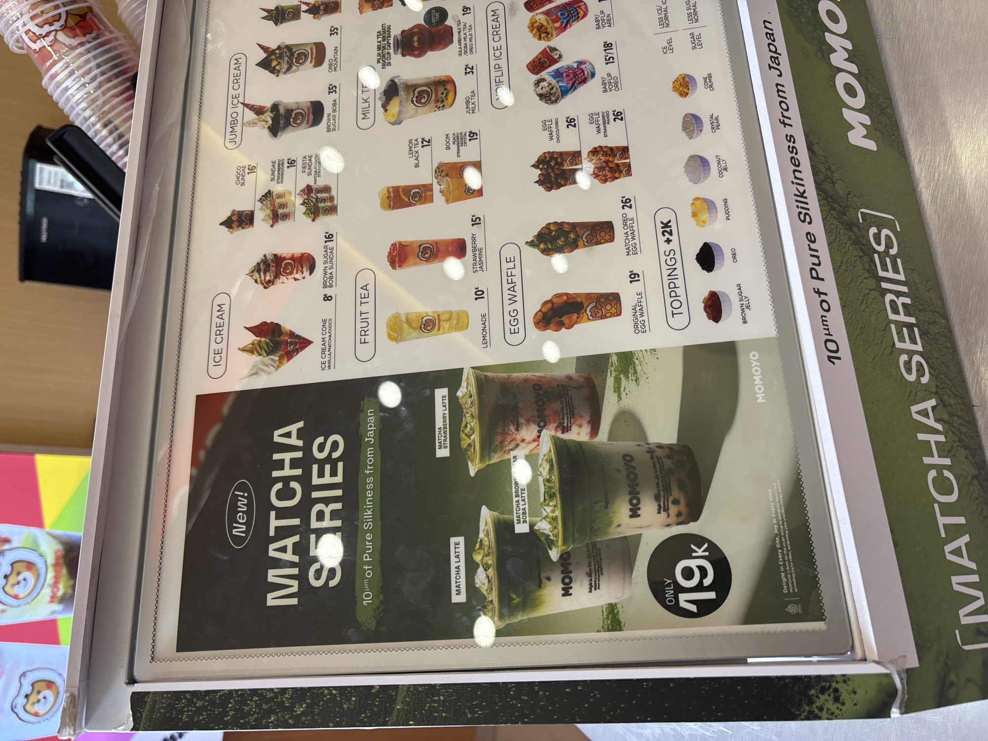 The image shows a menu board for "MOMOYO" a brand specializing in matcha-flavored desserts and drinks, photographed from a slightly skewed angle. The menu highlights the "MATCHA SERIES," featuring "10µm of Pure Silkiness from Japan," alongside other offerings such as Ice Cream, Fruit Tea, Jumbo Ice Cream, Yo!Flip Ice Cream, and Egg Waffles. The prices are displayed in Indonesian Rupiah (K = thousands), with the Matcha Latte priced at 19K. Toppings are available for an additional 2K. The menu also lists various drink flavors and toppings, with specific ice and sugar level options. The location is likely in North Jakarta, Indonesia, based on the brand's presence and the use of Indonesian currency. Several plastic cups are visible to the side, suggesting the menu is displayed in the store.