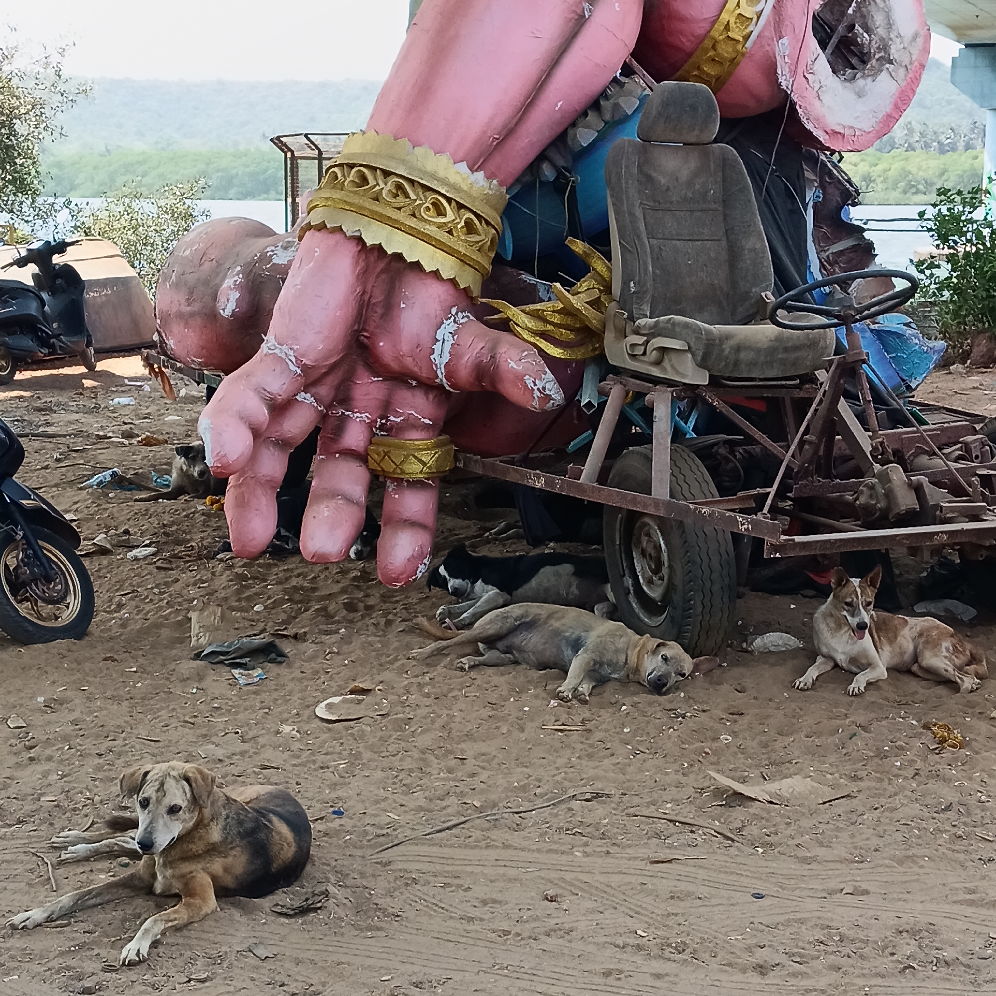 The image depicts a sandy area in Chopdem, India, littered with trash. Several stray dogs are resting on the ground. In the background, a large, disused sculpture of a hand with gold rings is visible, attached to a trailer with an old car seat and steering wheel. Two motorcycles are parked nearby. A body of water with green vegetation is in the distance.