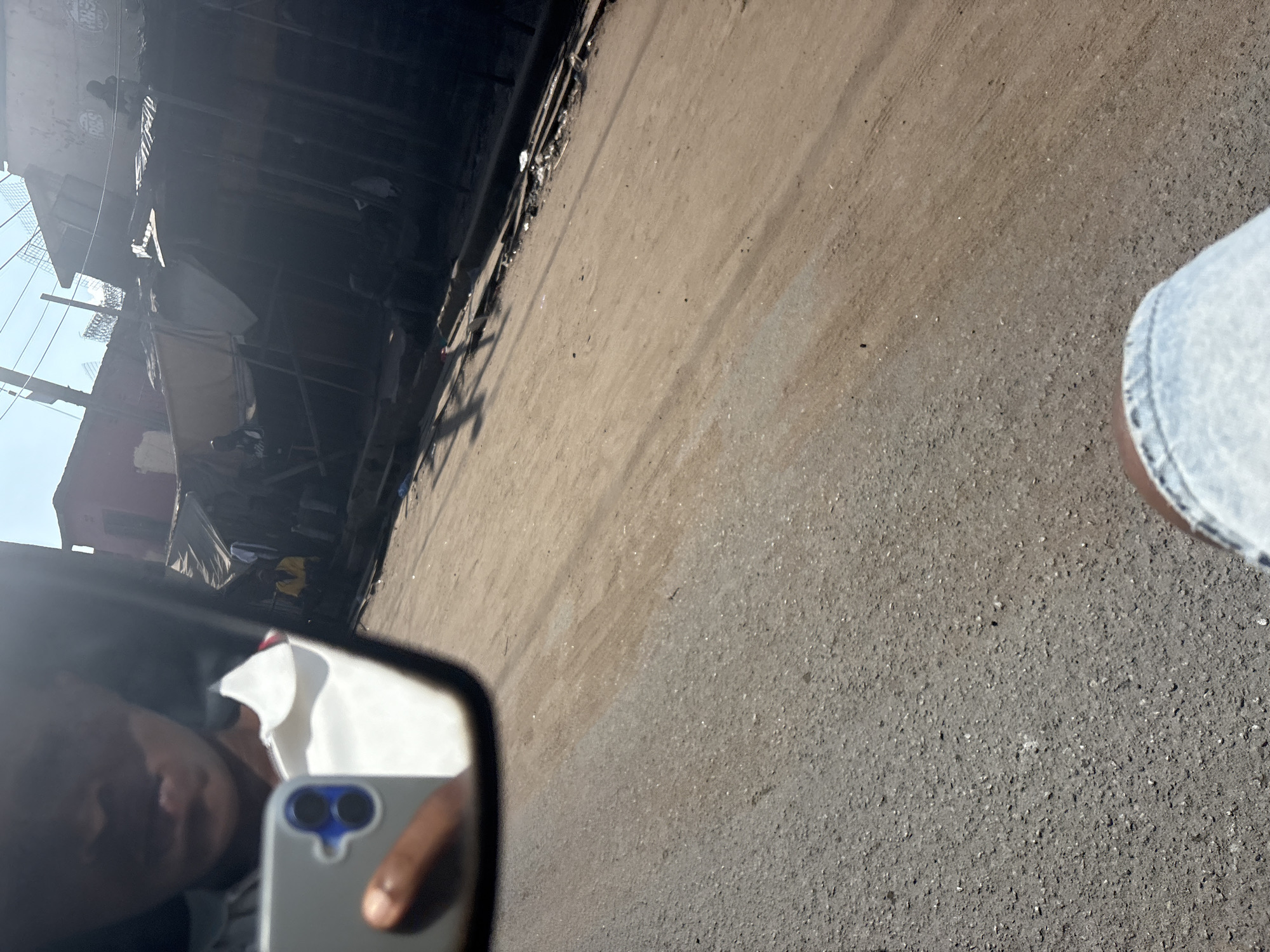 The image is a reflection in a car mirror, showing a person holding a smartphone. The reflection captures the person's face, a portion of their white shirt, and the smartphone. In the background, there's a street scene in Fegge, Nigeria. The street appears to be unpaved and dusty, with market stalls and buildings lining one side. Several people are visible in the background, some standing and others sitting. The overall impression is of a busy, outdoor marketplace.
