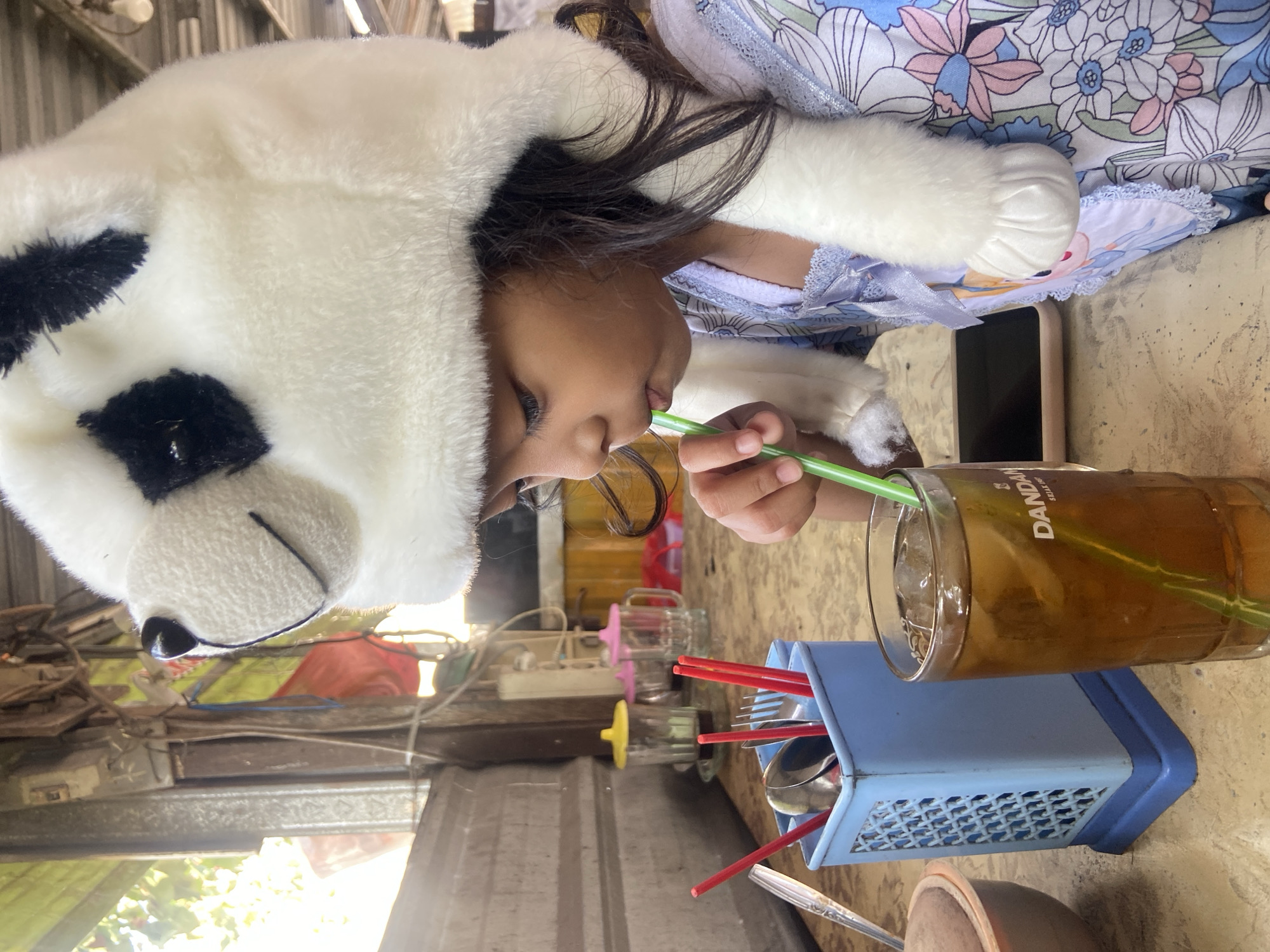 In this image, a young child wearing a fluffy white and black panda hat with attached paws is drinking from a glass of iced tea with a green straw. The child's face is partially obscured by the hat, but their eyes are visible, and they appear to be concentrating on drinking. The child is wearing a light blue and white floral-patterned garment.
The scene is set at a table, likely outdoors, given the dappled light and the background which includes some rustic structures and greenery. A blue plastic utensil holder with red chopsticks and silverware is visible next to the glass. A smartphone is also placed on the table. The overall setting suggests a casual, possibly informal, environment. The presence of the panda hat and the bright lighting hint at a daytime setting.
The visible text on the glass reads "DANDANG" and below it "SULTAN RAJA". The image has a warm tone, suggesting it was taken on a bright day. The specific location context is Balonggabus, Indonesia. The child's focused expression while drinking implies a moment of simple enjoyment or perhaps a break from an activity.