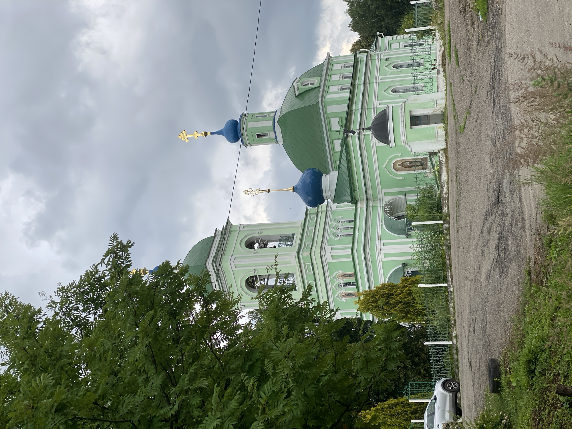 An exterior, eye-level, full shot captures a light green Orthodox church with a bell tower. The church has a large, green, domed roof, and a smaller blue dome topping the bell tower, also with a green domed roof. Multiple golden crosses are visible atop the domes. The church's walls are a pastel green with white trim and arched windows. A small, dark, dome-shaped structure is attached to the side of the church. Green trees are to the left of the church, and a green metal fence runs in front of it, with a silver car parked behind it. The ground in front of the church is paved with asphalt, and there is a spare tire lying on the ground. The sky is cloudy and overcast.