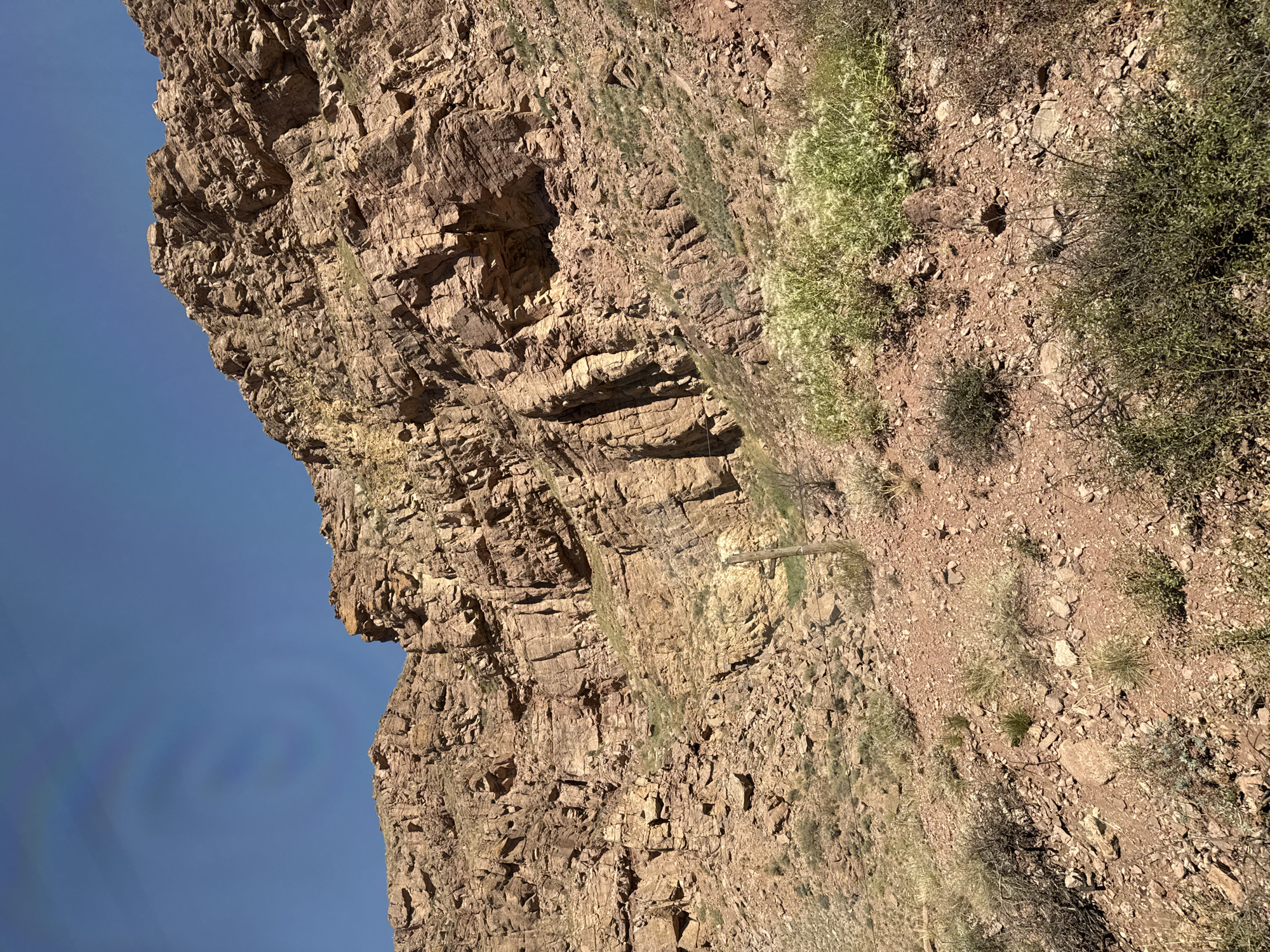 The image captures a rocky mountainside under a clear blue sky. The terrain is characterized by layered sedimentary rock in shades of brown and reddish-brown, with visible striations and fissures. Sparse vegetation, including dry grasses and low-lying shrubs with small white flowers, grows on the slopes and at the base of the mountain. A weathered wooden post with wire fencing is present on the lower slope. The overall environment appears arid and natural, suggesting a remote or undeveloped area. The location is described as the city of Krasnyy Most, Kyrgyzstan.