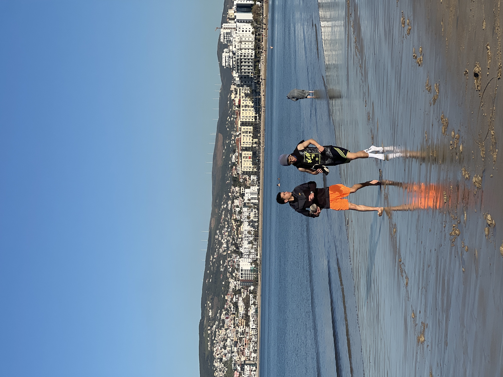 Two young men stand in the wet sand of a beach, reflecting in the water. One is wearing a dark jacket and bright orange shorts, holding a black object in one hand. The other wears a black top with yellow details, black shorts, white socks, and a gray hat, also holding a black object. A person in dark clothing stands further out in the water. Behind them, a cityscape rises along a hill with densely packed buildings and wind turbines on the horizon. The sky is clear blue. The location appears to be a beach in the city of Tanger, Morocco.