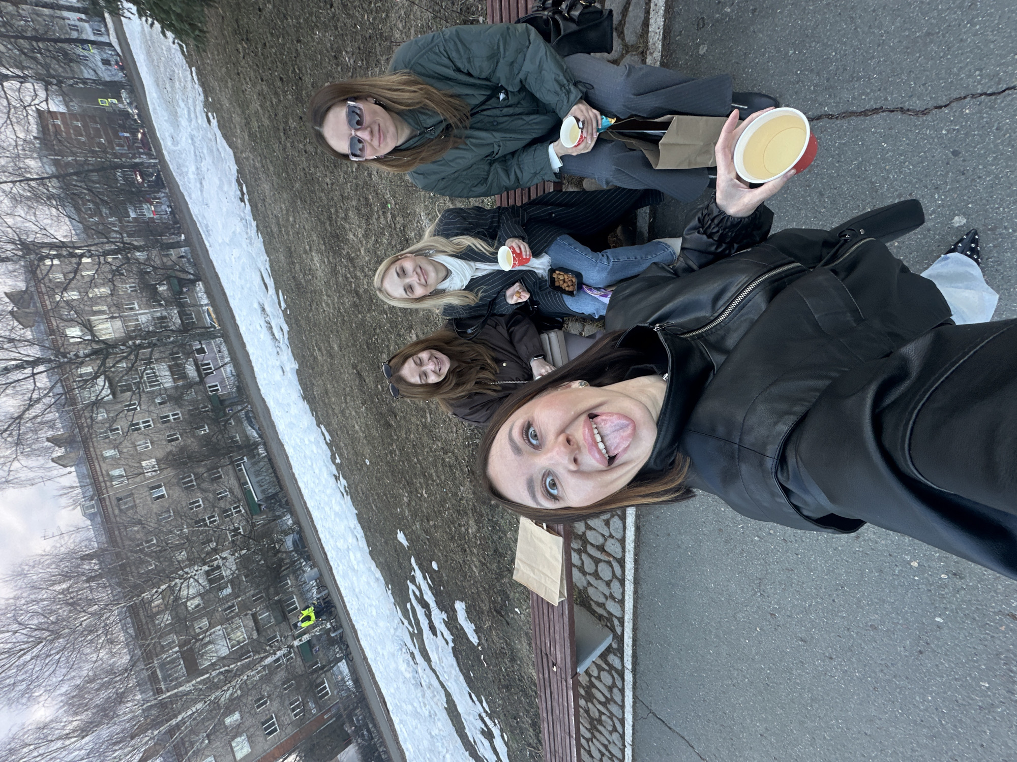 Four young women are sitting on a park bench. They are dressed in jackets and appear to be drinking something, possibly coffee or tea. The woman closest to the camera is taking a selfie and sticking her tongue out. There is snow on the ground near the bench. In the background, there are buildings, implying an urban setting. The location is Izhevsk, Russia.
