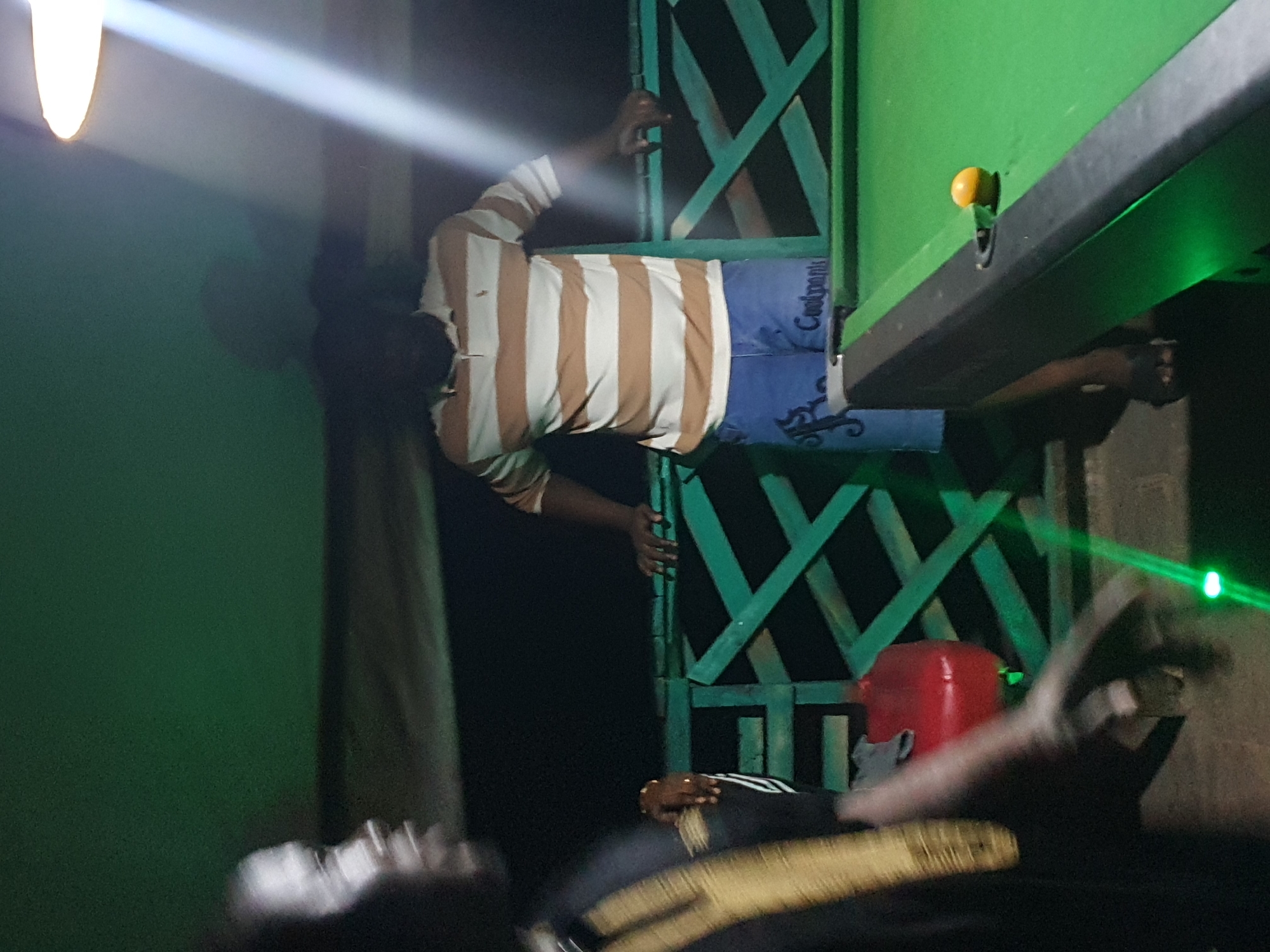 This is a medium-shot, eye-level, outdoor photograph taken at night in Lokoja, Nigeria. The scene appears to be a recreational area, possibly a bar or a private gathering space, featuring a pool table. A man in the foreground is partially visible on the left, with his face turned away from the camera, his body angled towards the pool table. He is wearing a dark shirt with a gold-colored stripe across his chest. His hands are blurred, suggesting movement.
In the background, another man is standing next to a pool table. He is wearing a horizontally striped polo shirt in brown and white, and blue denim shorts. His left arm is resting on the railing, and his right hand is also visible. The pool table is green, with a single yellow ball positioned near the edge. A bright, white light source, possibly a lamp, illuminates the upper right portion of the image, casting a strong beam and creating shadows on the wall behind the man in the striped shirt. The environment includes a green wooden railing, a red spherical object, and what appears to be a dark, unpaved ground surface. The overall lighting is dim, with the exception of the strong overhead light.