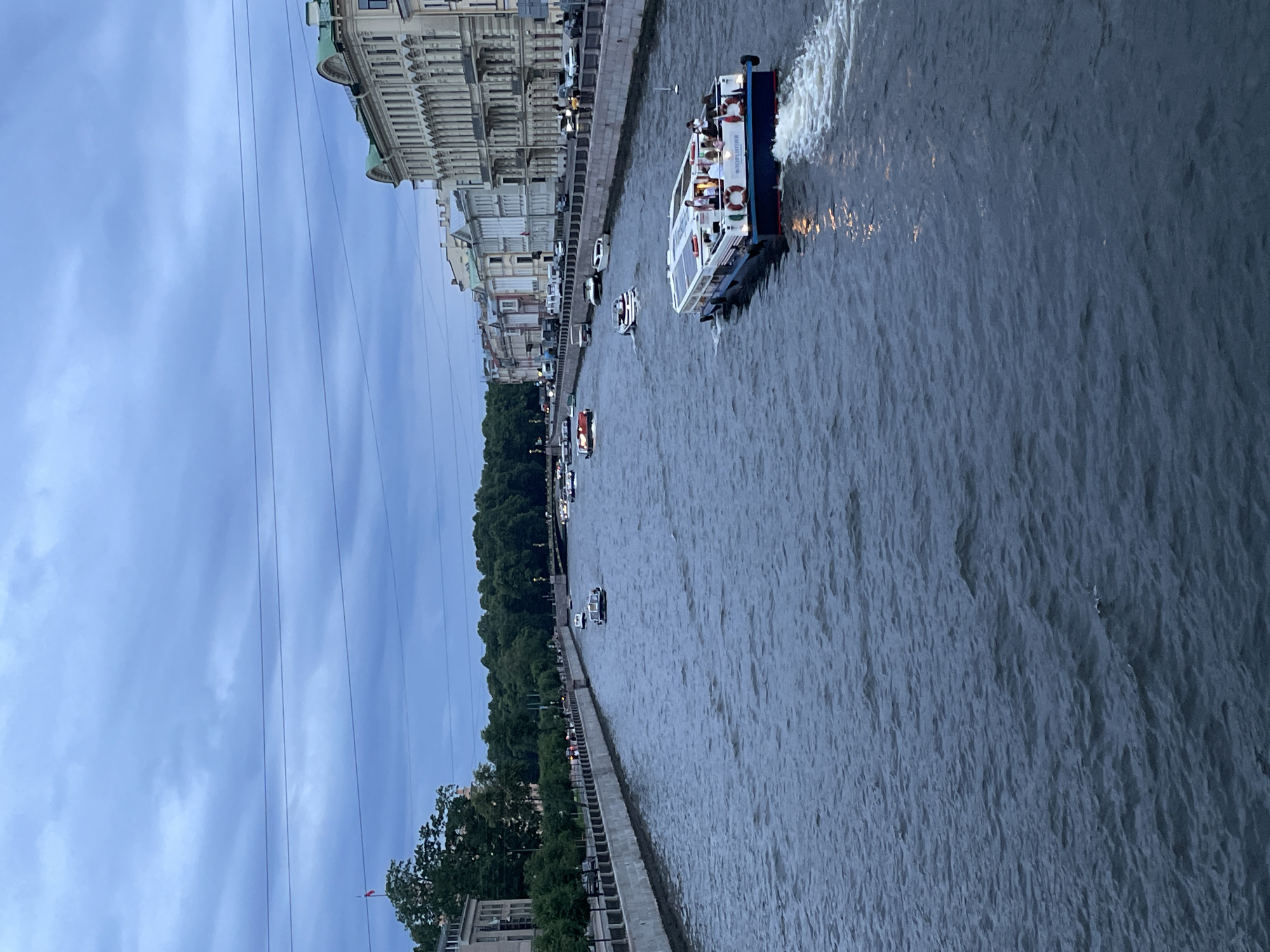 This is a vertical, eye-level, medium-shot photograph of the Griboyedov Canal in Saint Petersburg, Russia, taken on a cloudy day. A large, white tour boat with a blue stripe and people on its upper deck is moving away from the viewer towards the right, creating a white wake in the dark blue water. Several smaller boats are scattered further up the canal, some also appearing to be tour boats. The canal is lined on both sides by stone embankments. On the right bank, classical multi-story buildings with green roofs and arched windows are visible. On the left bank, there are trees lining the embankment, with some buildings and trees visible in the distance. Overhead power lines are visible against the cloudy sky. The overall atmosphere is calm and serene, typical of a European city with waterways.