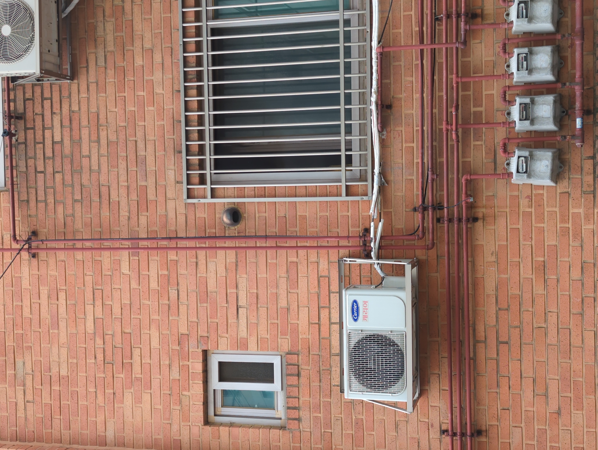 The image shows the exterior wall of a brick building. An air conditioning unit is mounted on a metal bracket on the wall to the left of a barred window. Below the air conditioning unit, a series of red pipes run horizontally across the brick wall, branching out vertically in some places. Four gas meters are attached to the wall along the bottom of the frame, connected to the red piping system. A smaller window with a white frame is visible above the air conditioning unit. The brickwork is a light orange-red color.