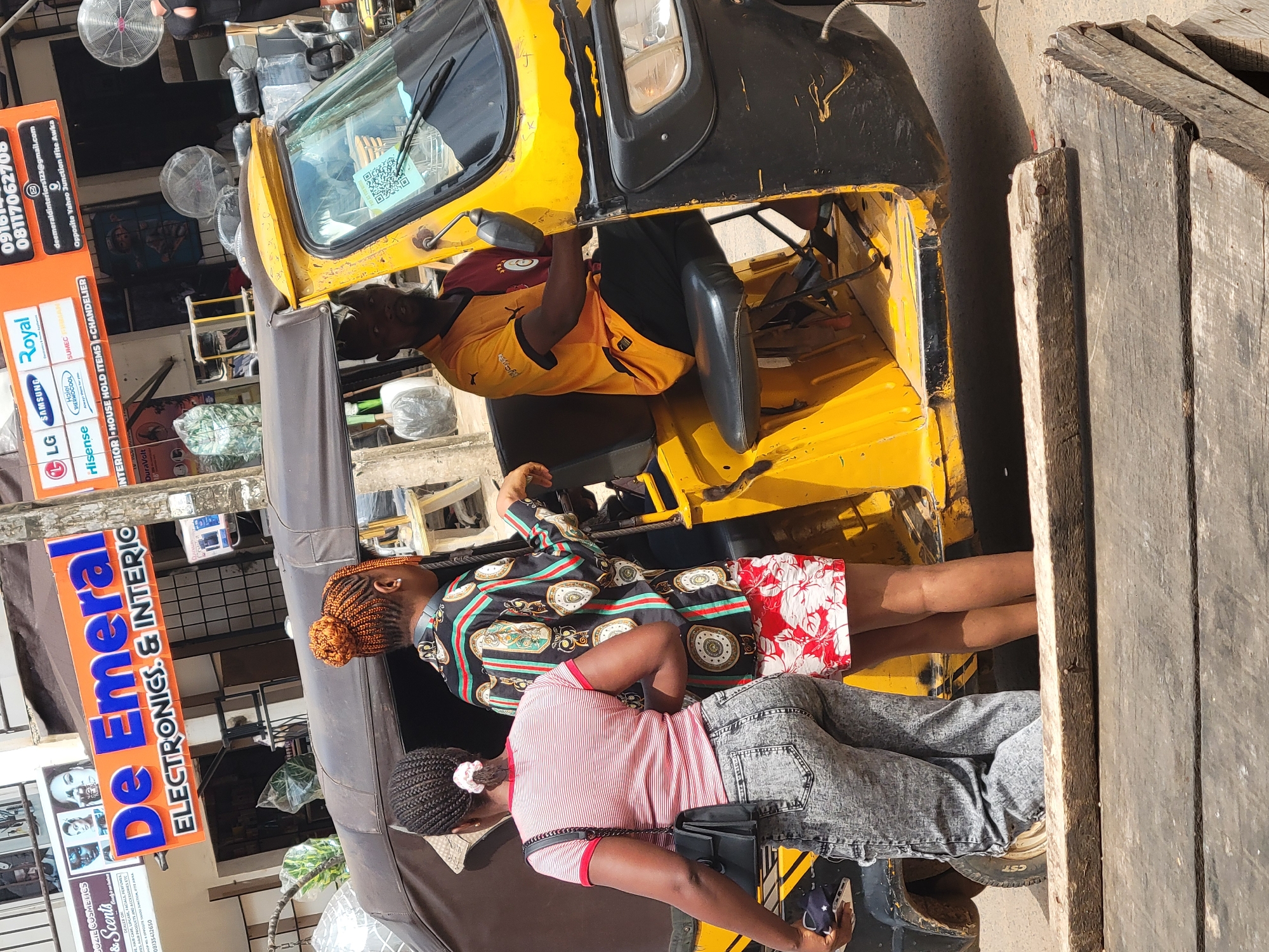 This image, taken in Egbagu, Nigeria, captures a street scene with a yellow and black auto-rickshaw as the central object. Two young women are visible in the foreground, seated within the open passenger area of the rickshaw. The woman on the left wears a patterned dress with gold, black, and red motifs, and her hair is styled in a bun. The woman on the right is dressed in a striped red and white t-shirt and denim shorts, with her hair in braids.
The rickshaw appears to be parked. A man, wearing a yellow and maroon shirt, is partially visible inside the driver's compartment, seemingly working on something or resting. A large, orange and blue sign for "De Emeral ELECTRONICS. & INTERIO" is prominently displayed on the left side of the frame, advertising electronics, interior design, house hold items, and chandeliers. Below this, smaller signs for brands like LG, Samsung, Hisense, and Royal are visible. A QR code is also displayed on the windshield of the rickshaw.
The scene suggests a daytime setting, with bright sunlight casting shadows. The environment appears to be a commercial area, given the shop signage. The overall mood is relaxed, with the individuals seemingly engaged in their daily activities. The presence of the auto-rickshaw and the street-level businesses indicates a bustling urban or semi-urban environment.