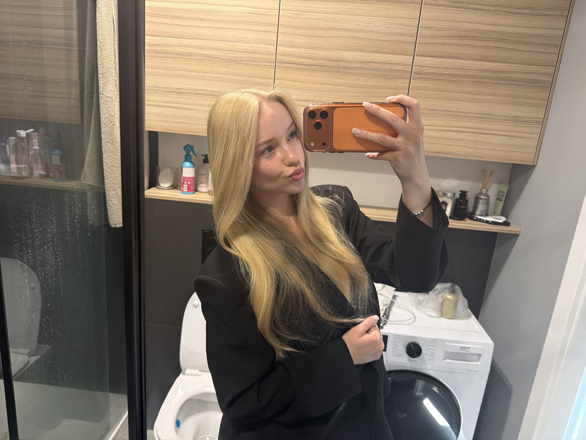 A young, light-skinned woman with long blonde hair is taking a selfie in a bathroom. She is wearing a black blazer. In the background, there is a toilet, a washing machine, wooden cabinets, and a glass shower. The location is in the city of Unknown, Unknown.