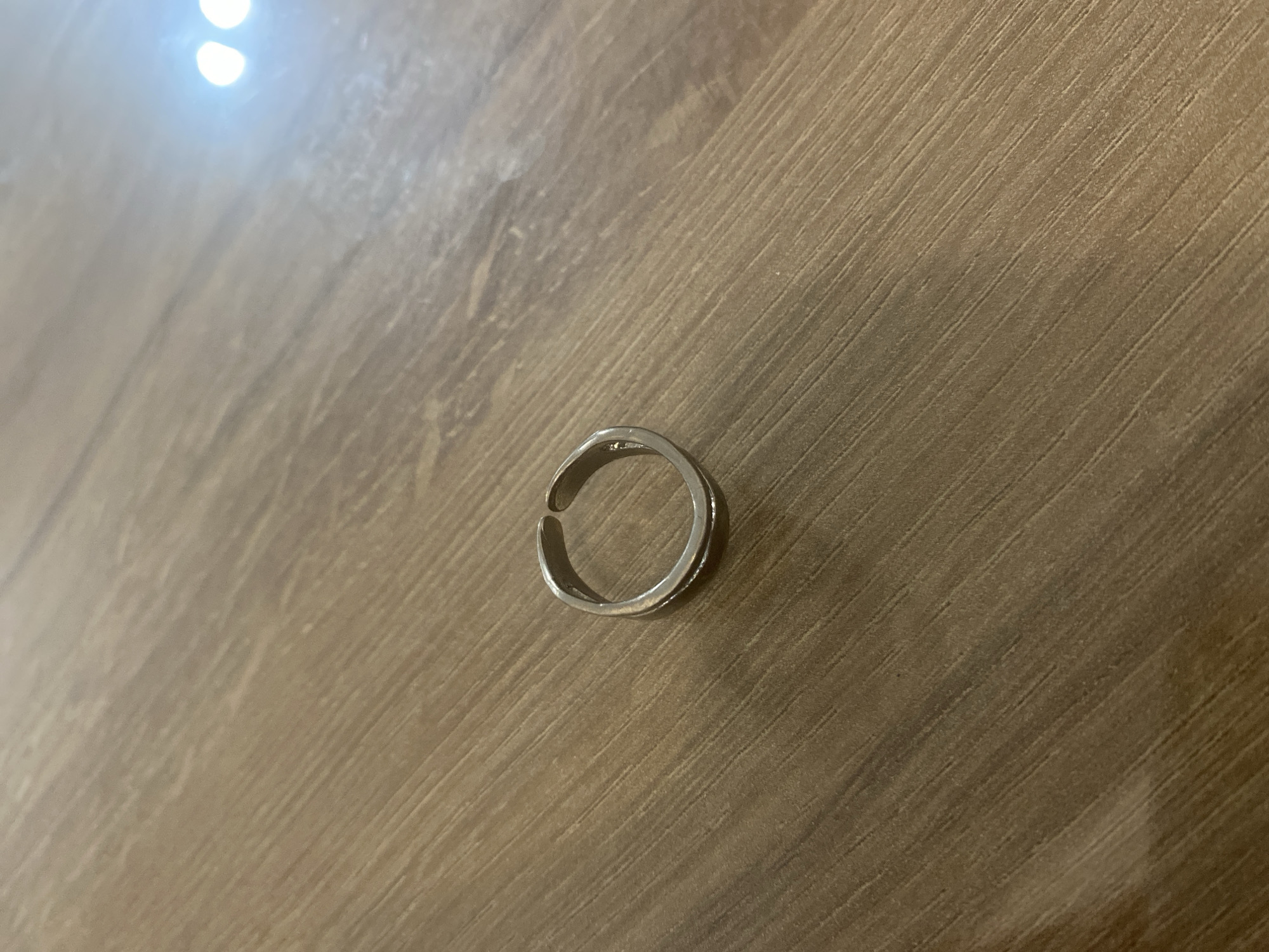 A silver-colored, open-ended ring is centrally positioned on a light brown wooden surface. The ring appears to be metallic and has a slightly textured or worn finish, with visible highlights reflecting light. The wooden surface exhibits a distinct linear grain pattern, running diagonally from the bottom-left to the top-right of the frame. Bright, circular reflections from an artificial light source are prominent in the upper-left area of the image, indicating an indoor setting. No people or activities are visible. There is no discernible text in the image.