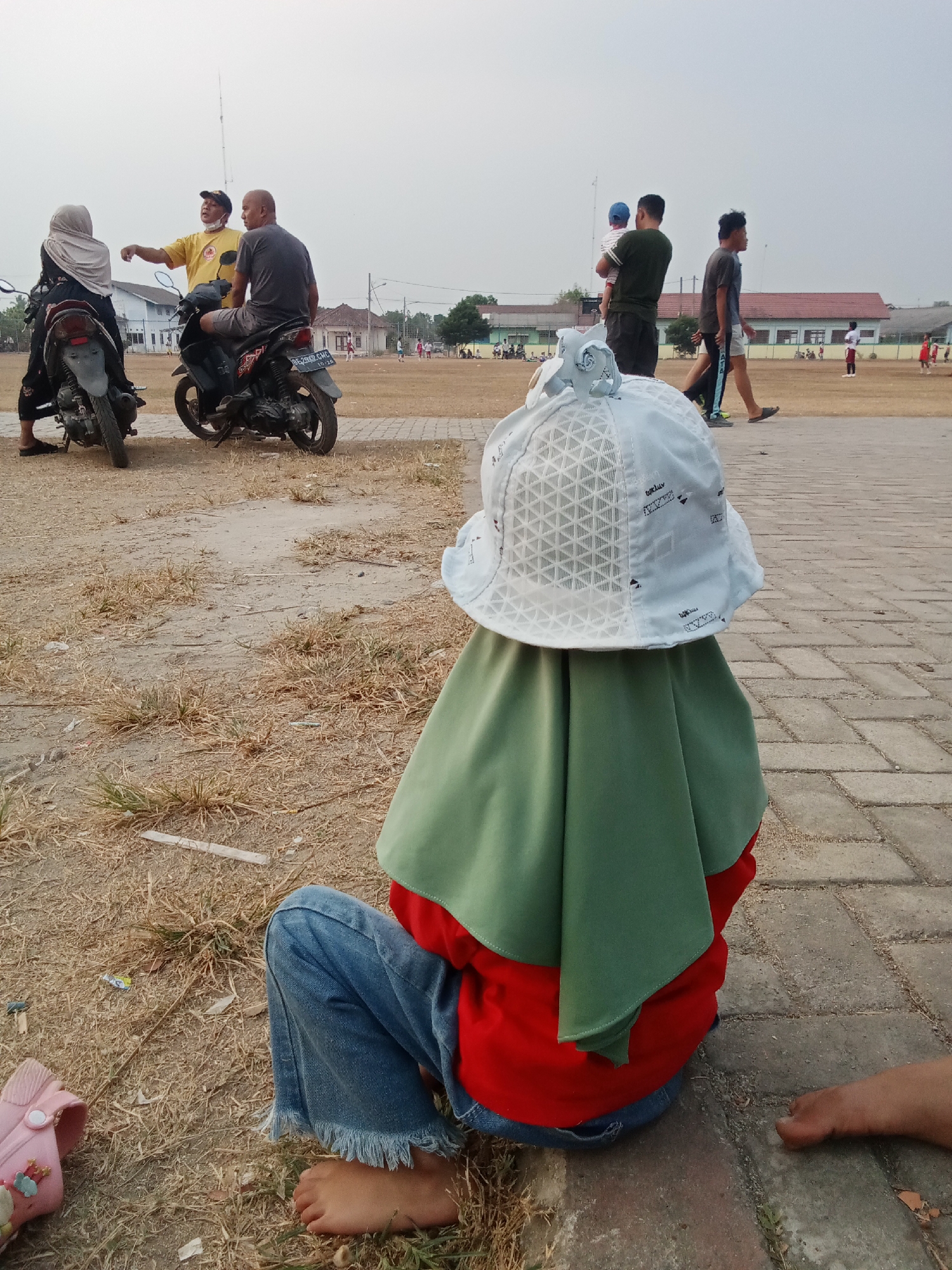 The image depicts a casual outdoor scene in Toto Katon, Indonesia, possibly a sports field or park. In the foreground, a child is seated, facing away from the camera, wearing a red shirt, blue jeans with frayed hems, and a light blue hat with a geometric pattern, partially obscuring their face with a green headscarf. The child is barefoot on a paved path next to a dry, grassy area.
In the mid-ground, two men and a woman are on motorcycles. One man, wearing a yellow shirt and a cap, gestures while speaking to another man in a gray t-shirt sitting on a motorcycle. The woman in a headscarf is seated behind the first man on his motorcycle. In the background, several people are visible, including a man holding a child, two men walking, and others engaged in activities on a field. Buildings with red roofs are present in the distance. The sky is overcast and pale.