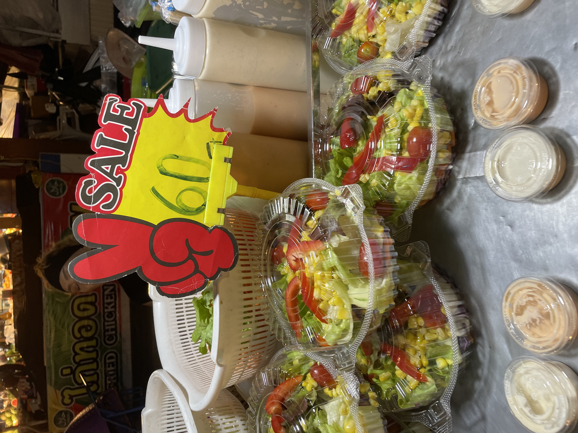 A street food stall in Pattaya City, Thailand, displays individual servings of salad in clear plastic containers. A red sign with a peace sign graphic and the word "SALE" in English and Thai script is prominently displayed, along with the price "60." Several bottles of salad dressing are lined up behind the salads. In the background, a person is partially visible behind the signage, and the stall appears to be set up for business with various food items and equipment. The overall scene depicts a busy market environment.