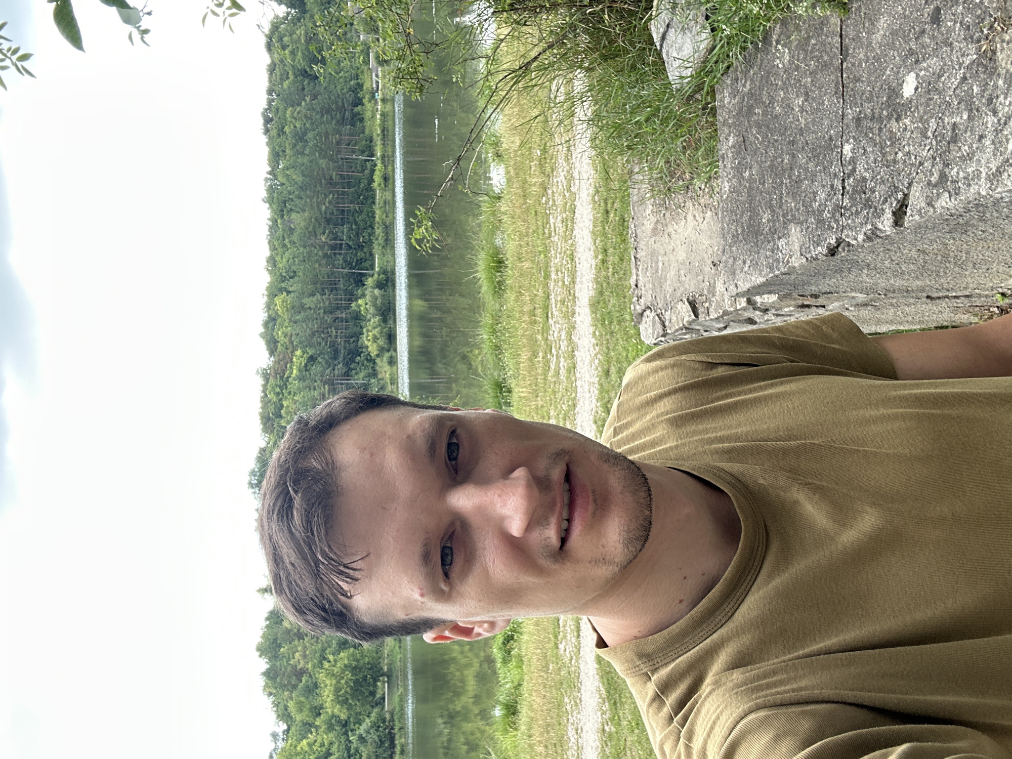 A young man with short brown hair and a light beard is taking a selfie in front of a lake and a forest. He is wearing a tan t-shirt. The man is in the foreground, and the lake and forest are in the background. The sky is cloudy. The scene is located in Makiv, Ukraine.