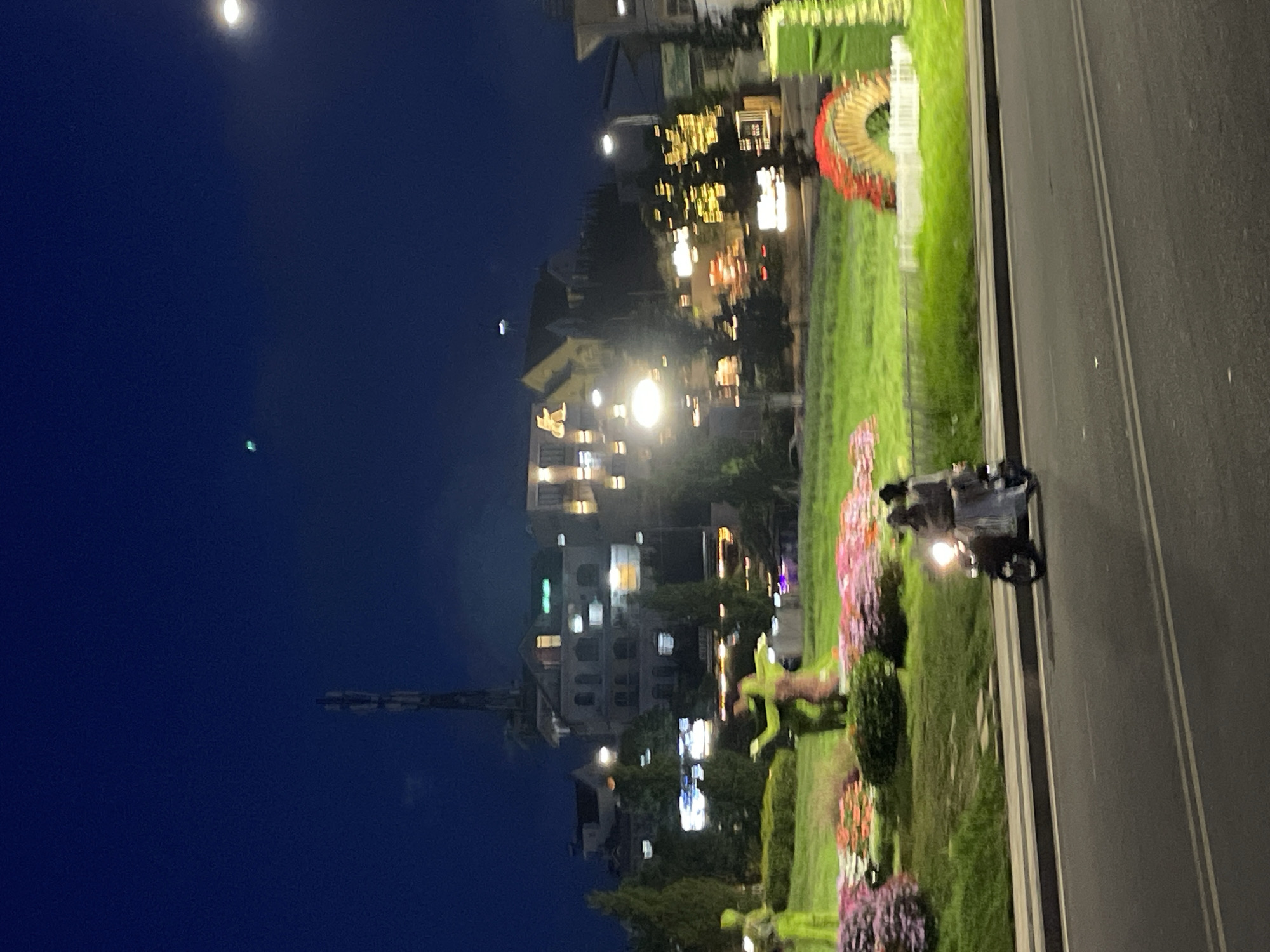 This is a nighttime photograph depicting an urban scene in Da Lat, Vietnam, characterized by a mix of natural elements, artificial light, and architectural structures.
In the foreground, a dark asphalt road with two faint parallel white lines extends across the bottom of the frame. To the right of the road, a concrete curb and sidewalk separate it from a lush green park area.
In the midground, the park features expansive green grass punctuated by colorful flower beds in shades of pink, purple, and orange-red. Several sculptures are visible within the park: two prominent, stylized human-like figures, one in neon green and another in a muted green/brown, appear to be interacting or dancing. Another neon green figure stands further to the left. To the right, decorative structures include a red and gold arched element and a vertical green panel. Sparse, conical trees are also present in the park.
Behind the park, various buildings with illuminated windows and signs form the city's backdrop. A communication tower, tall and slender, rises prominently against the dark sky on the left. On a building towards the right, a large, illuminated yellow "K" sign is clearly visible. Numerous smaller lights from buildings and streetlights dot the scene, creating a diffused glow.
In motion on the road in the foreground, two individuals are riding a scooter, traveling from right to left. The scooter's bright white headlight is illuminated. The front rider wears a dark top, while the passenger is dressed in a light-colored long garment and a dark top.
The sky is a deep, dark blue, indicative of night. A single faint green light source is visible high in the sky to the upper left of the "K" sign, and another bright, circular light source (likely a distant streetlight or moon) is present in the upper right corner. The image exhibits a slight softness and blur, consistent with low-light photography or minor camera movement.