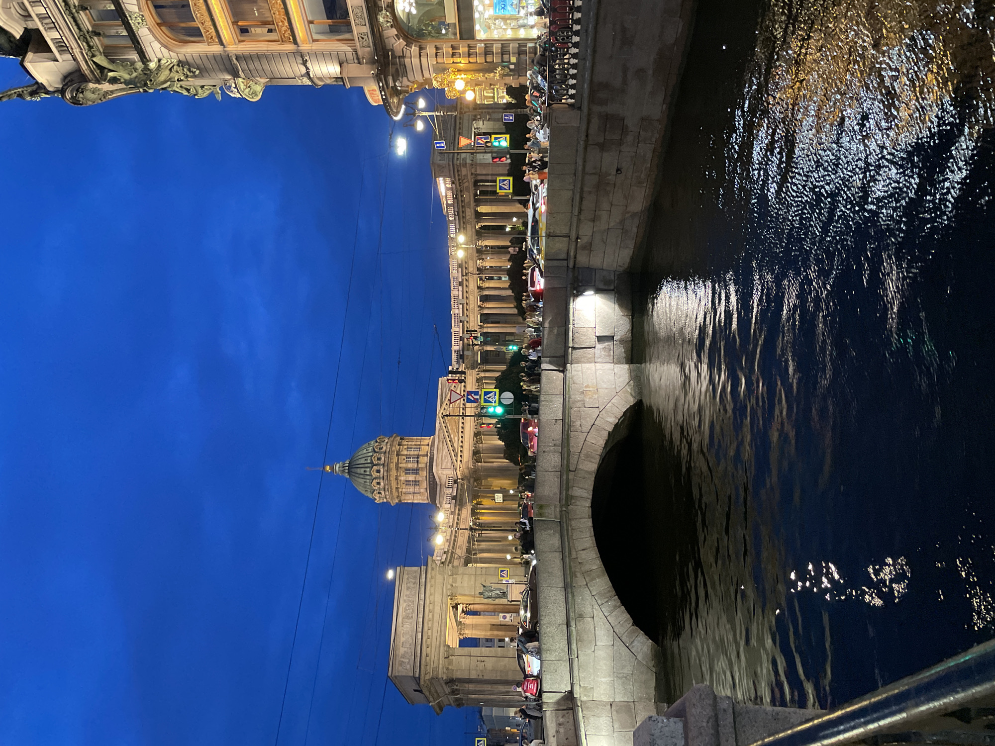 This is a night-time, vertical, medium-shot photograph of the Kazan Cathedral in Saint Petersburg, Russia, viewed from a bridge over a canal. The sky is a deep, dark blue, with hints of lighter blue and clouds. The cathedral, with its iconic green dome and colonnade, is illuminated and dominates the background. To the right of the cathedral, a building with an ornate facade and illuminated windows is visible. In the foreground, a stone bridge arches over a dark canal. The water reflects the lights from the buildings and streetlights, creating shimmering patterns on the surface. Several cars and pedestrians are present on the bridge and sidewalk. Traffic lights and street signs are also visible. The overall atmosphere is one of a bustling city scene at dusk.