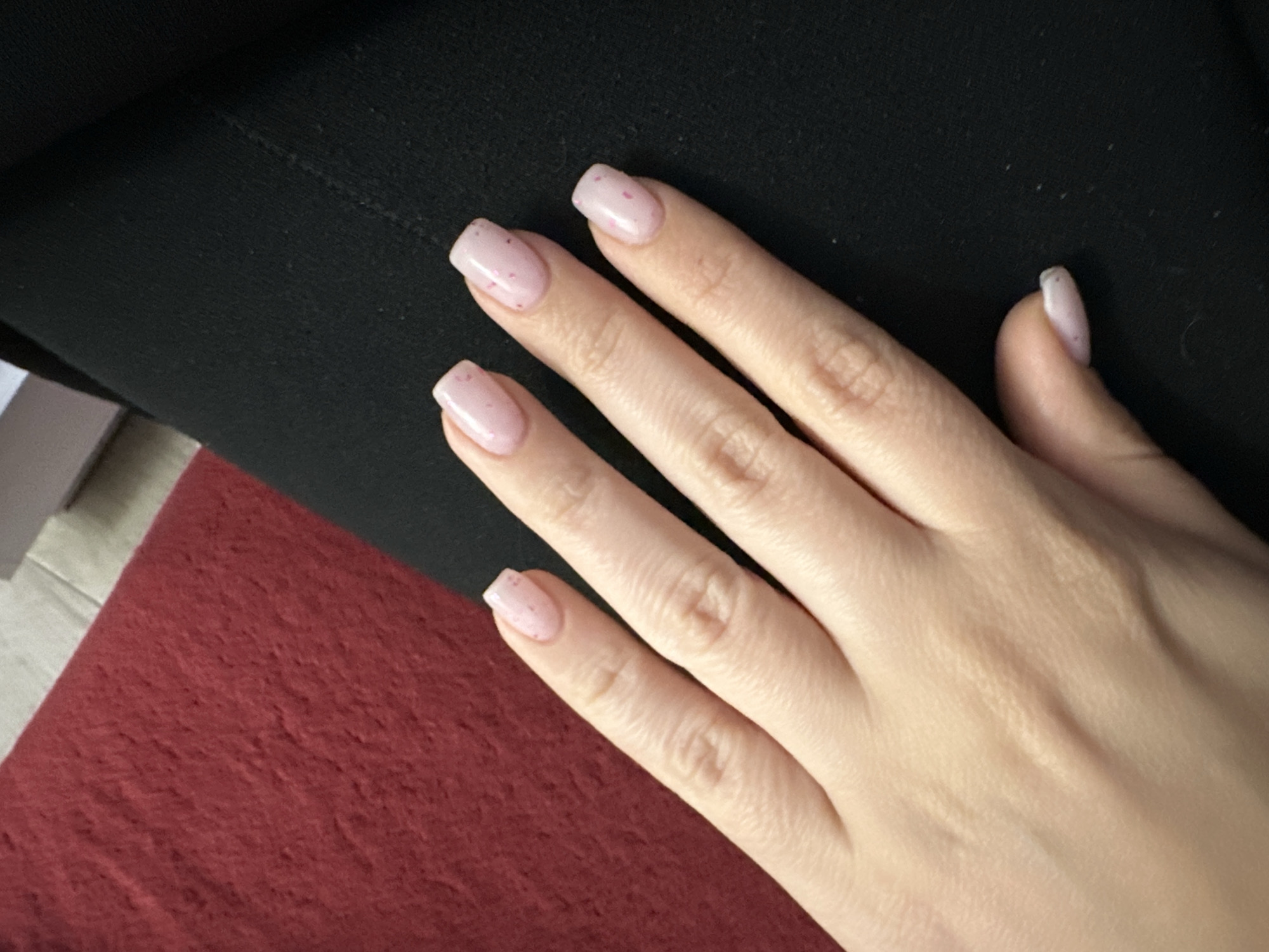 A human hand with painted fingernails lies on a black surface. The fingernails are painted in a light pink color with sparse pink speckles. Part of a dark red surface is visible in the lower left corner. A gray surface is visible on the left edge of the image, adjacent to the red surface. The scene is indoors, and the location of Sofia, Bulgaria cannot be determined from the visual information provided.