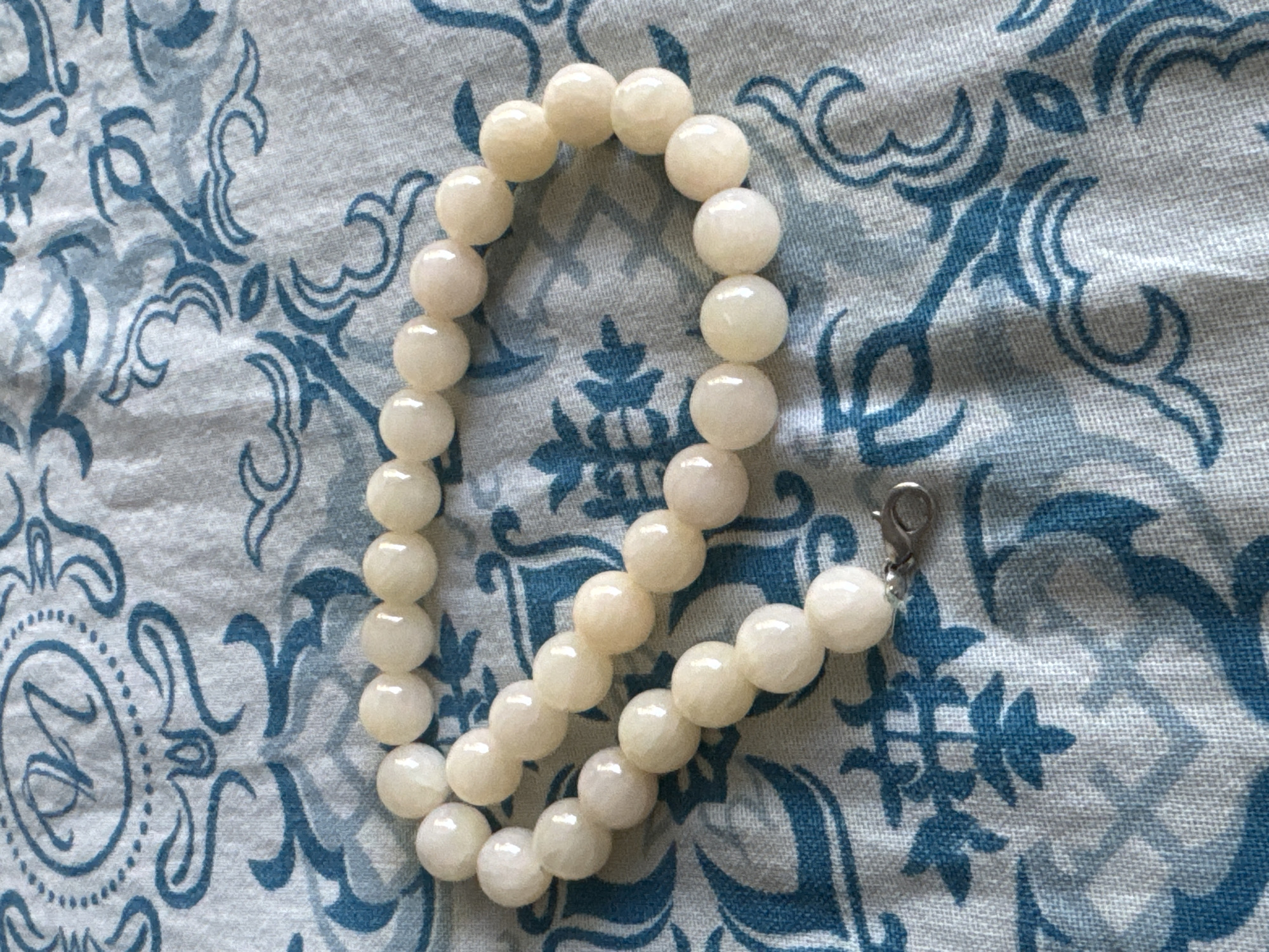A close-up, eye-level shot shows a strand of pale, creamy-white beads with a smooth, polished finish. The beads are spherical and vary slightly in size, strung together on a fine silver chain. A silver clasp, designed in a lobster-claw style, is visible towards the bottom left of the frame, connecting the ends of the strand. The beads are arranged in a loose, curved pattern against a light blue fabric with a white damask-like pattern. The pattern on the fabric features stylized floral or abstract motifs in a darker shade of blue, creating a visually contrasting background. There are no people or animals present in the image. The setting appears to be indoors, on a surface covered by the patterned fabric. The lighting is even, with no harsh shadows, suggesting ambient light. The image does not provide any information about a specific location in Яромінскі сельскі Савет, Belarus, or any other geographical context.