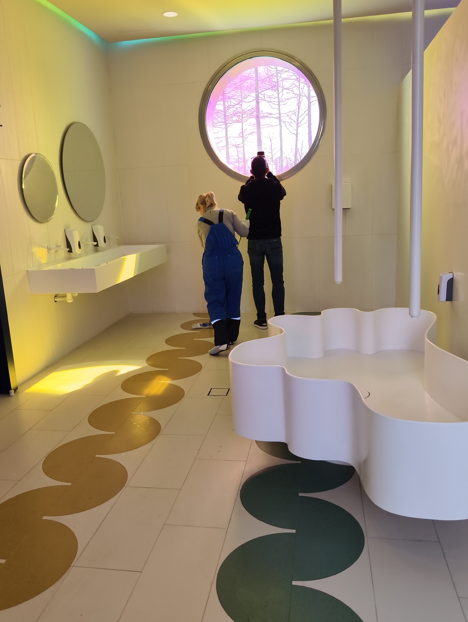 The image captures a vibrant, modern interior space, likely a public restroom or an artistic installation, located in Krasnodar, Russia. The room is characterized by its contemporary design and dynamic lighting.
Two individuals are visible. On the left, a person, possibly a woman, dressed in blue overalls and a light-colored top, is bending down, appearing to attend to something on the floor. Her blonde hair is tied back. In the center-right, a man wearing a dark long-sleeved shirt and jeans stands facing away from the viewer. He is looking out of a large, circular window with a metallic frame and holding what appears to be a phone or camera to take a picture of the scene outside.
The interior walls are white, complemented by striking overhead lighting that casts colorful gradients of yellow, green, and orange across the walls and floor. A long, minimalist white sink counter with multiple faucets and hand soap dispensers is mounted on the left wall, above which hang two oval mirrors. The floor is tiled in a light color, featuring large, sinuous golden-brown circular patterns that weave across the space, leading towards a section with dark green circular patterns. On the far right, a unique, low-lying white basin with a wavy, organic shape adds to the distinctive aesthetic. Two slim white poles extend vertically from the ceiling towards the right side of the room.
Through the circular window, bare tree branches are visible, suggesting an autumn or winter season. The light filtering through the window is tinted with a purple/pink hue, creating a surreal outdoor view. The scene conveys a sense of modern design and artistic flair, with the activities of the individuals adding a human element to the visually engaging environment. No legible text is visible in the image.