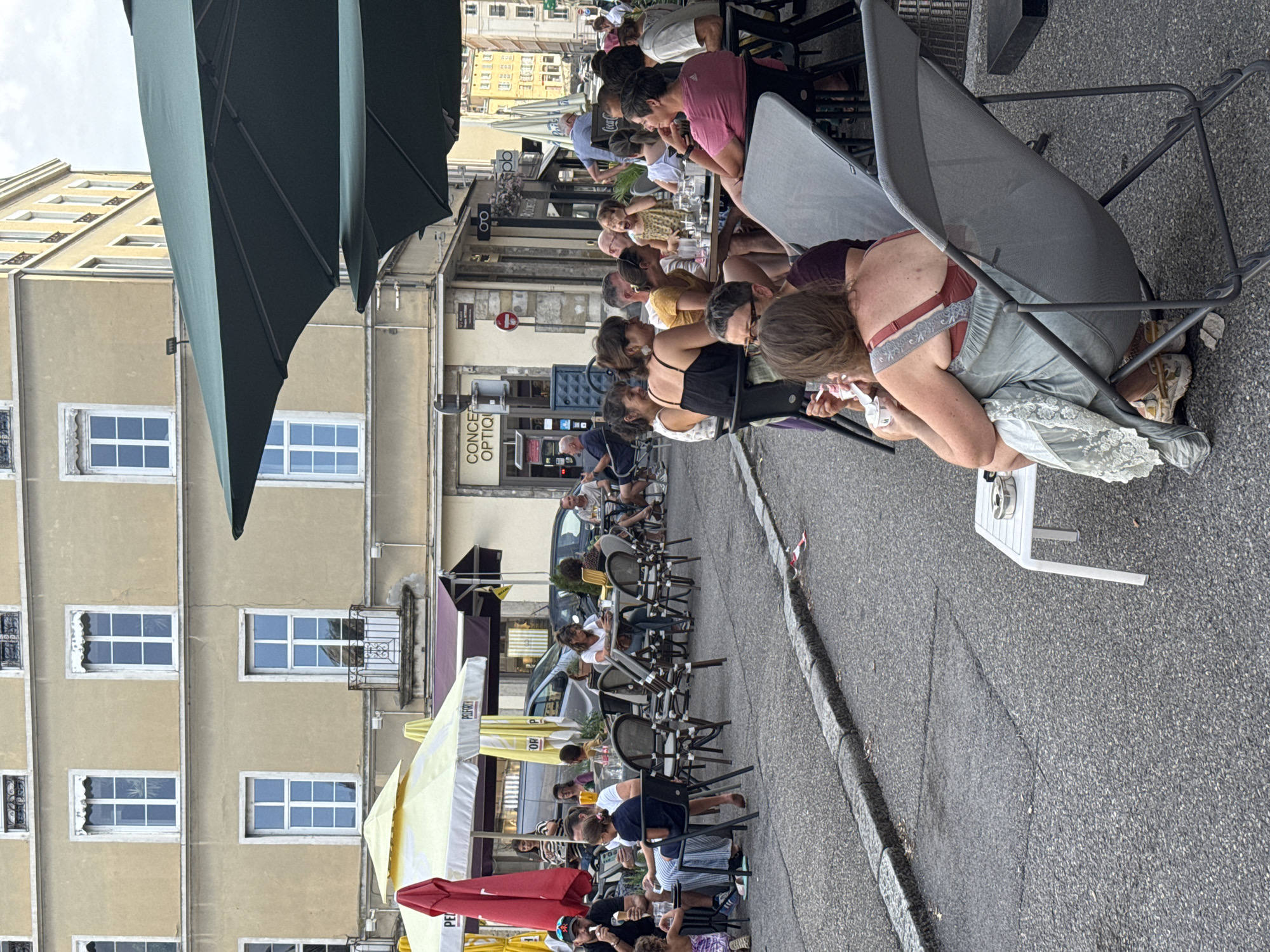 The image depicts an outdoor seating area of a cafe or restaurant on a city street in Vienne, France. Several groups of people are seated at tables, some eating or drinking and engaging in conversation. In the foreground, a woman sits in a gray lounge chair, wearing a light blue dress with lace trim and a reddish-brown bra. She is holding a container and appears to be eating ice cream. To her left, a small white table holds an ashtray. Behind her, other patrons are visible at various tables. The background shows a building with multiple windows, likely an apartment building or shopfront, with awnings over some of the windows. Several parked cars are visible further down the street. Large green umbrellas are providing shade over some of the seating areas. The ground is paved with asphalt.