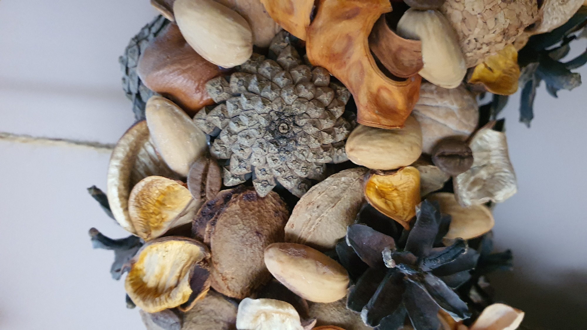 The image is a close-up, still life shot of a potpourri decoration. The potpourri is composed of various dried natural elements including pine cones, dried seed pods, nuts, and coffee beans. The arrangement is intricate and textured, with a variety of earthy tones and natural shapes. The background is a plain, light-colored wall, suggesting the decoration is displayed indoors. There are no people present in the image. The scene is static and does not depict any action.
There is no information available within the image or its crops to determine its location in Krasnoyarsk, Russia.