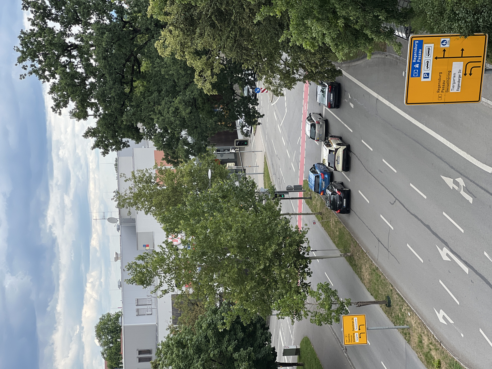The image is an aerial view of a street intersection in Straubing, Germany, on an overcast day. Several cars are present on the road, including a blue sedan, a white taxi, a silver SUV, and a black sedan. Traffic lights are visible, and a pedestrian crossing with red markings is present. Two yellow directional signs are posted, one indicating routes to "Regensburg", "Passau", and "Tiergarten", and another providing similar directions with additional options for "Geiselhörring" and "Theresiencenter". Tall green trees line the street and a modern building with a white facade is visible in the background. The sky is filled with clouds.