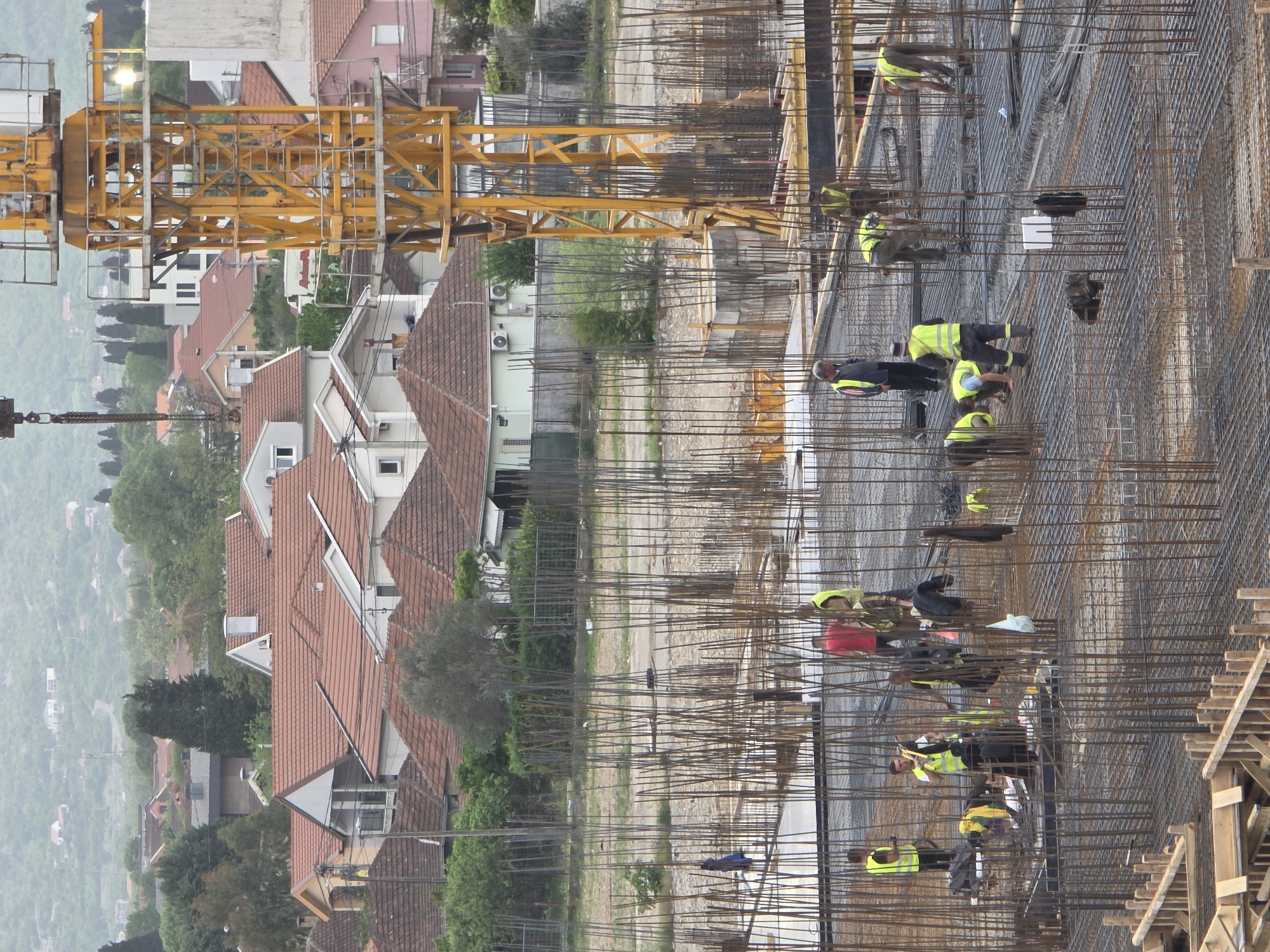 The image captures an active construction site in Podgorica, Montenegro, seen from an elevated perspective. In the foreground, a vast area is covered with a dense grid of steel reinforcement bars (rebar), indicating the initial stages of a concrete structure like a foundation or floor slab. Numerous construction workers are present, all wearing high-visibility yellow or green vests, diligently working among the rebar. They are seen bending over, likely involved in tying, positioning, or inspecting the steel framework.
A prominent yellow tower crane dominates the midground, its long horizontal jib extending across the scene, underscoring the scale of the construction project. Beyond the immediate construction zone, a residential neighborhood unfolds, characterized by houses with distinctive red-tiled roofs nestled among trees. In the background, a landscape of hazy, green hills or mountains stretches into the distance, suggesting an urban area bordering natural terrain. The sky appears overcast or hazy, contributing to a soft, diffused lighting across the scene. The image depicts a busy daytime work environment. No visible text is clearly legible on any objects or clothing.