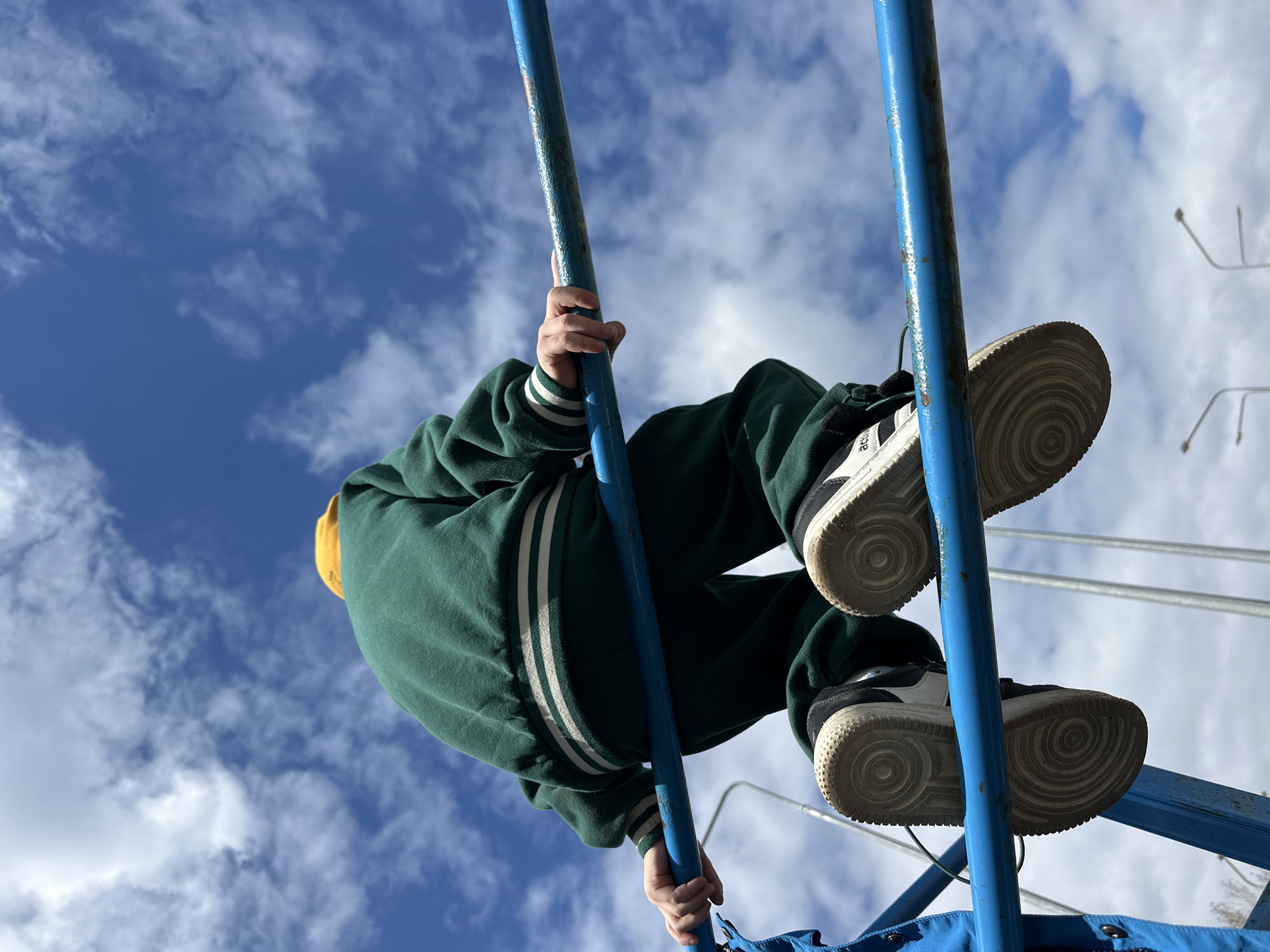 A person is hanging upside down from a blue metal structure against a partly cloudy sky. The person wears a green jacket with white stripes on the sleeves, black and white sneakers, and a yellow hat or hood. Their hands are gripping the metal bars. The structure appears to be part of playground equipment. The location is the city of Chaikovsky, Russia.