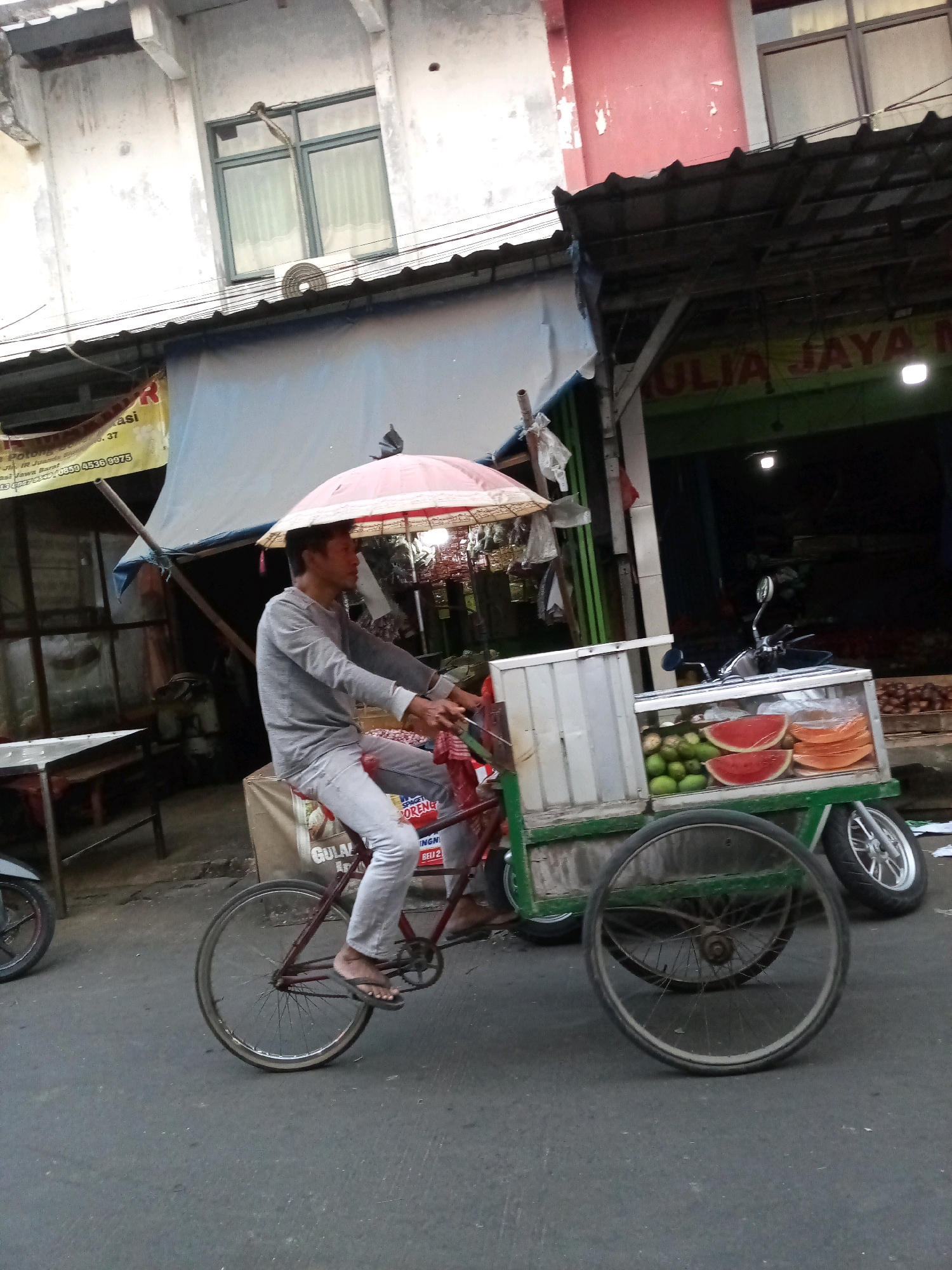 In Bekasi, Indonesia, a man rides a red bicycle equipped with a cart and an umbrella, navigating a street lined with shops. The cart, attached to the rear of the bicycle, is a mobile fruit stand displaying sliced watermelons and other fruits. The man, wearing a light gray long-sleeved shirt and light-colored pants, is pedaling the bicycle. He holds the handlebars with his right hand while his left hand rests near the cart. An umbrella shields him from the sun. In the background, shops with signs and goods are visible, indicating a bustling market or commercial area. The street is paved, and the overall scene suggests a typical day in a busy Indonesian city.