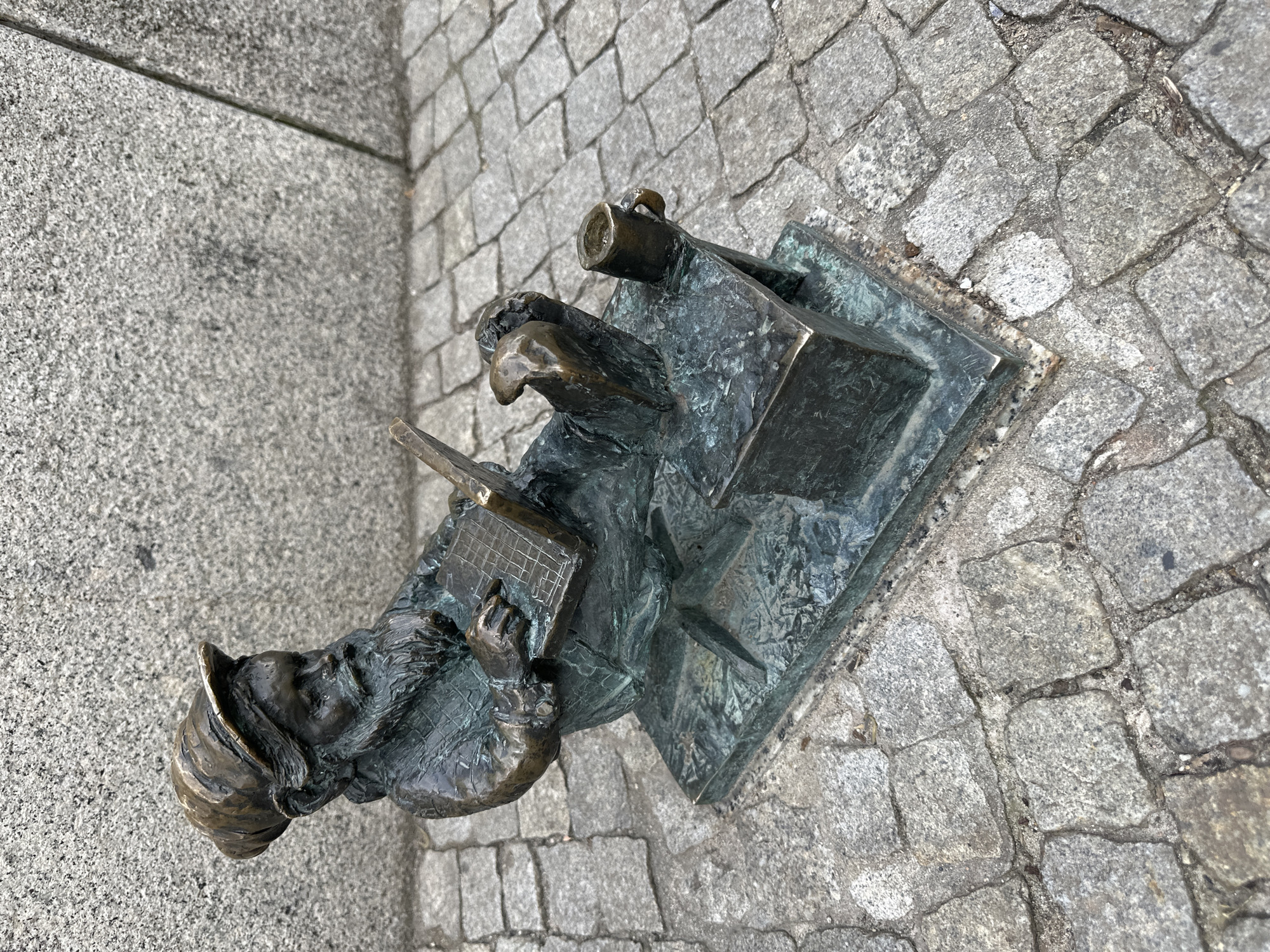 A small, bronze statue of a gnome stands on a stone-paved street. The gnome is seated at a desk with a quill in hand and a beer stein on the desk behind him. The gnome is wearing a hat and has a focused expression. The location is Wrocław, Poland, where numerous gnome statues are placed throughout the city.
