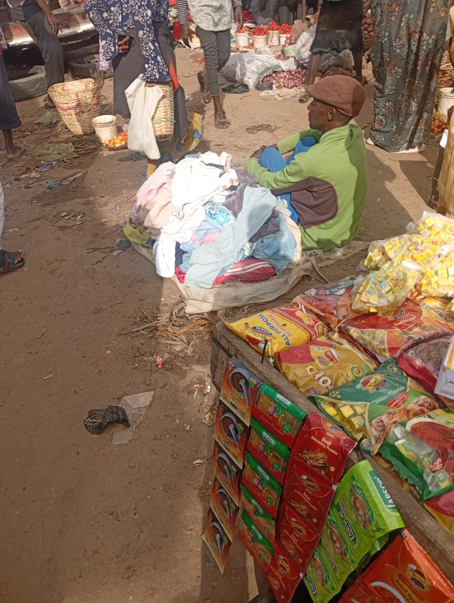 The image depicts an outdoor market scene in Rafin Par, Nigeria. In the foreground, a man wearing a green long-sleeved shirt and a brown cap sits on the ground, looking to his right. He is surrounded by piles of colorful clothing. To the right, a wooden stall is laden with various packaged food items, including cubes of seasoning (beef and chicken flavors), curry powder, and rice seasoning. Some packages are labeled "Mr. Chef," "Gino," and "LARSOR." In the background, several people are visible, some standing and others in the process of buying or selling goods. One person is seen near a basket filled with what appear to be fruits or vegetables. The ground is dusty and uneven, typical of an open-air market. The lighting suggests it is daytime, with sunlight casting shadows. The overall atmosphere appears to be one of commerce and daily life in a bustling market. Visible text includes brand names and product descriptions like "Beef Seasoning Powder," "Curry Powder," "Chicken Flavour," and "Jollof Rice."